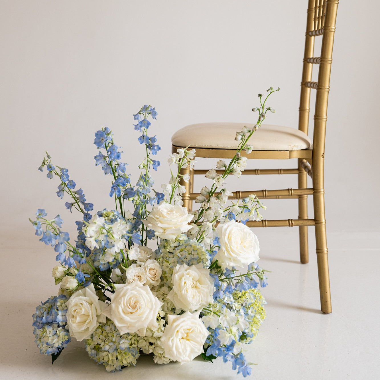 A white and light blue floral arrangement with white roses and blue delphiniums placed in front of a gold Chiavari chair against a plain off-white background.