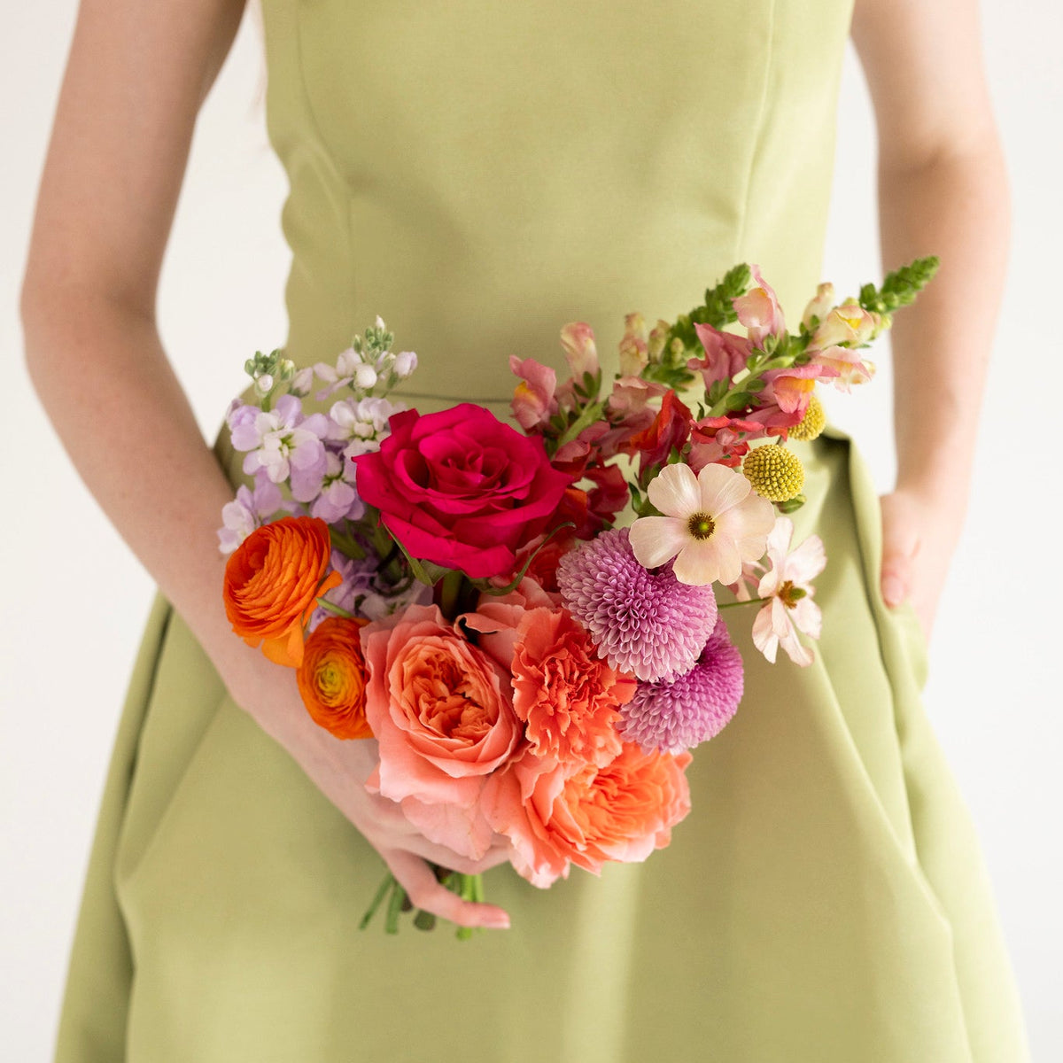 A person wearing a light olive green dress holds a vibrant, multicolored floral bouquet featuring pink, orange, purple, and white flowers against a clean white background.