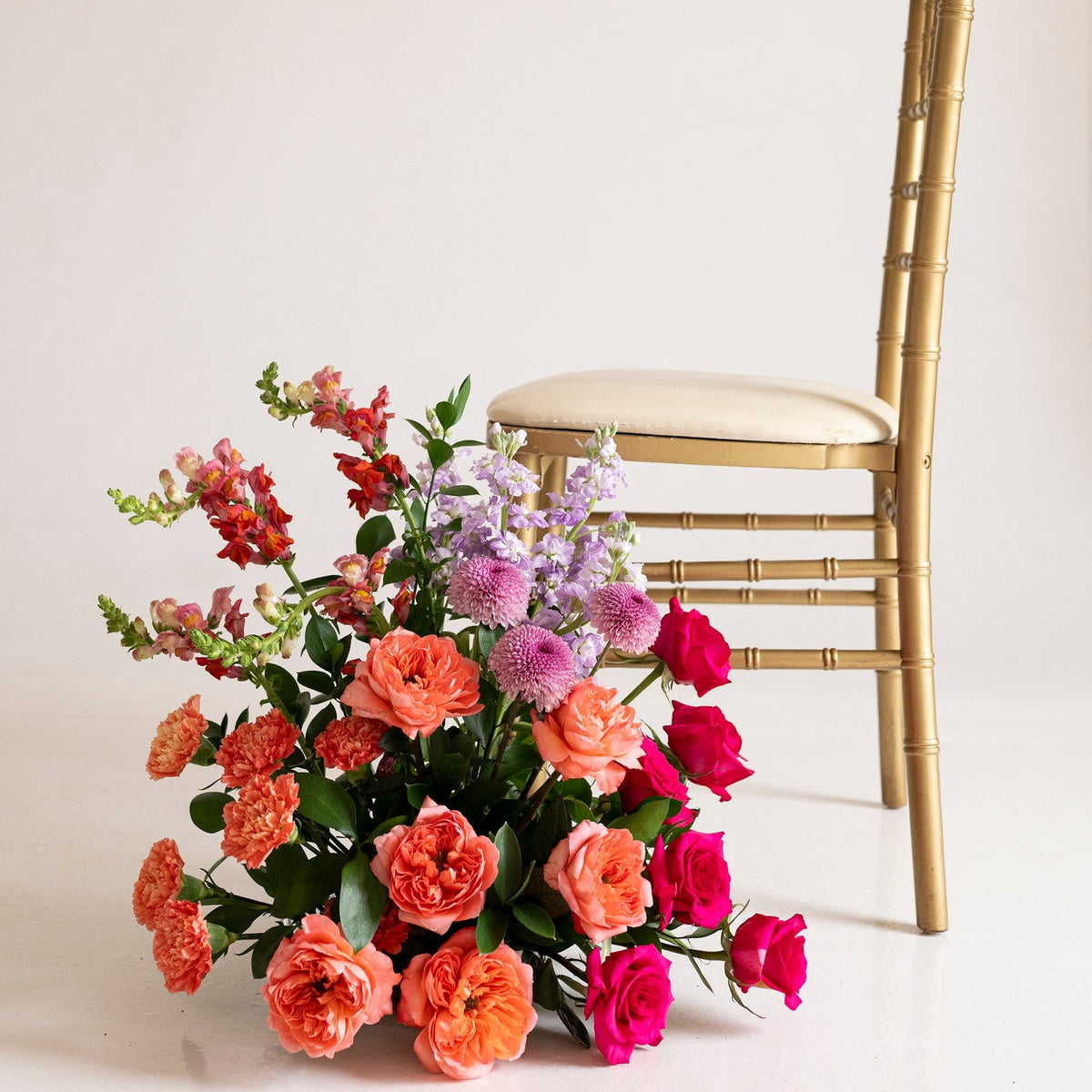 A vibrant floral arrangement of pink, red, and orange roses with green foliage and purple filler flowers, styled next to a gold bamboo-style chair against a clean white background.