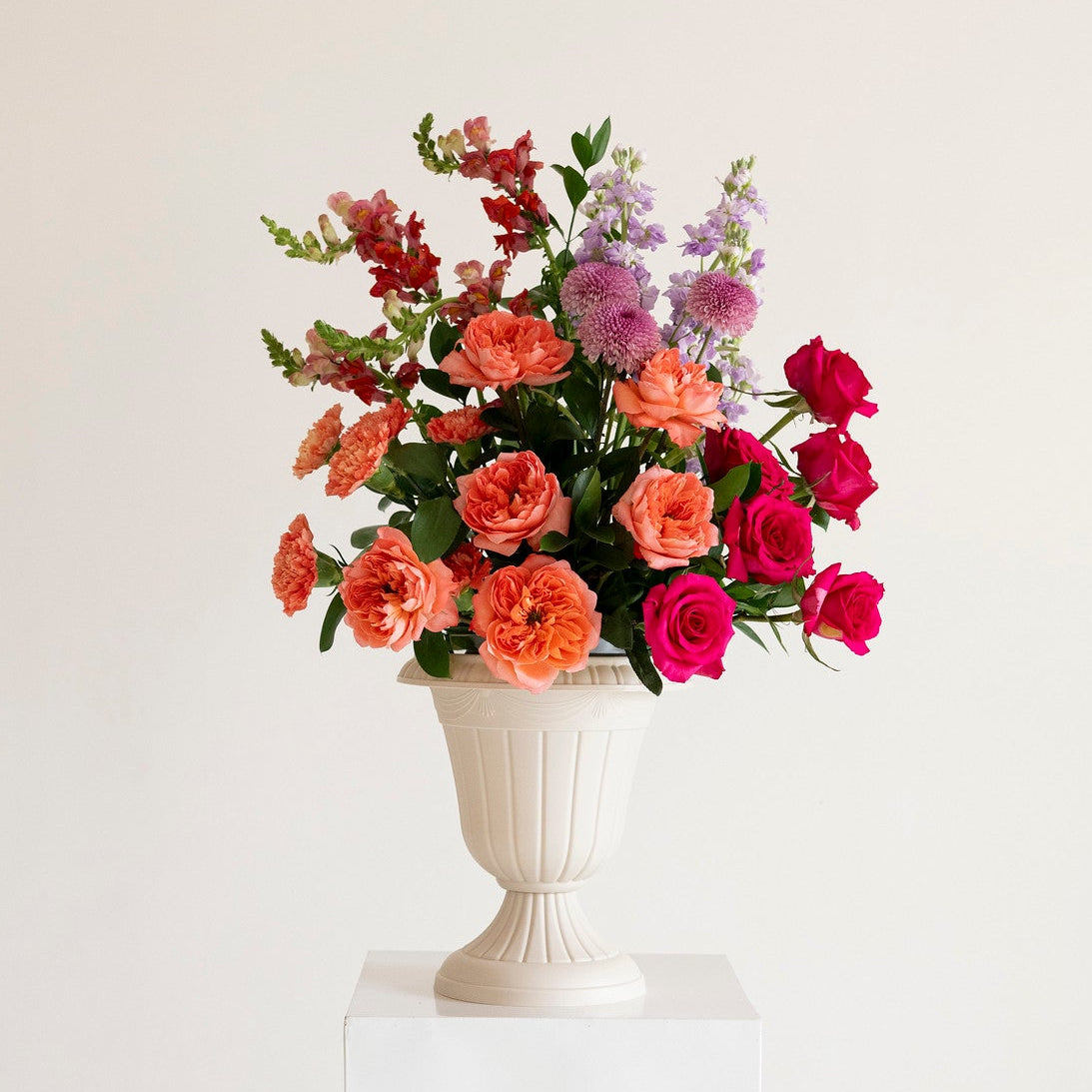 A vibrant fresh flower bouquet in a white classical-style urn vase, displayed on a white pedestal against a clean white background.