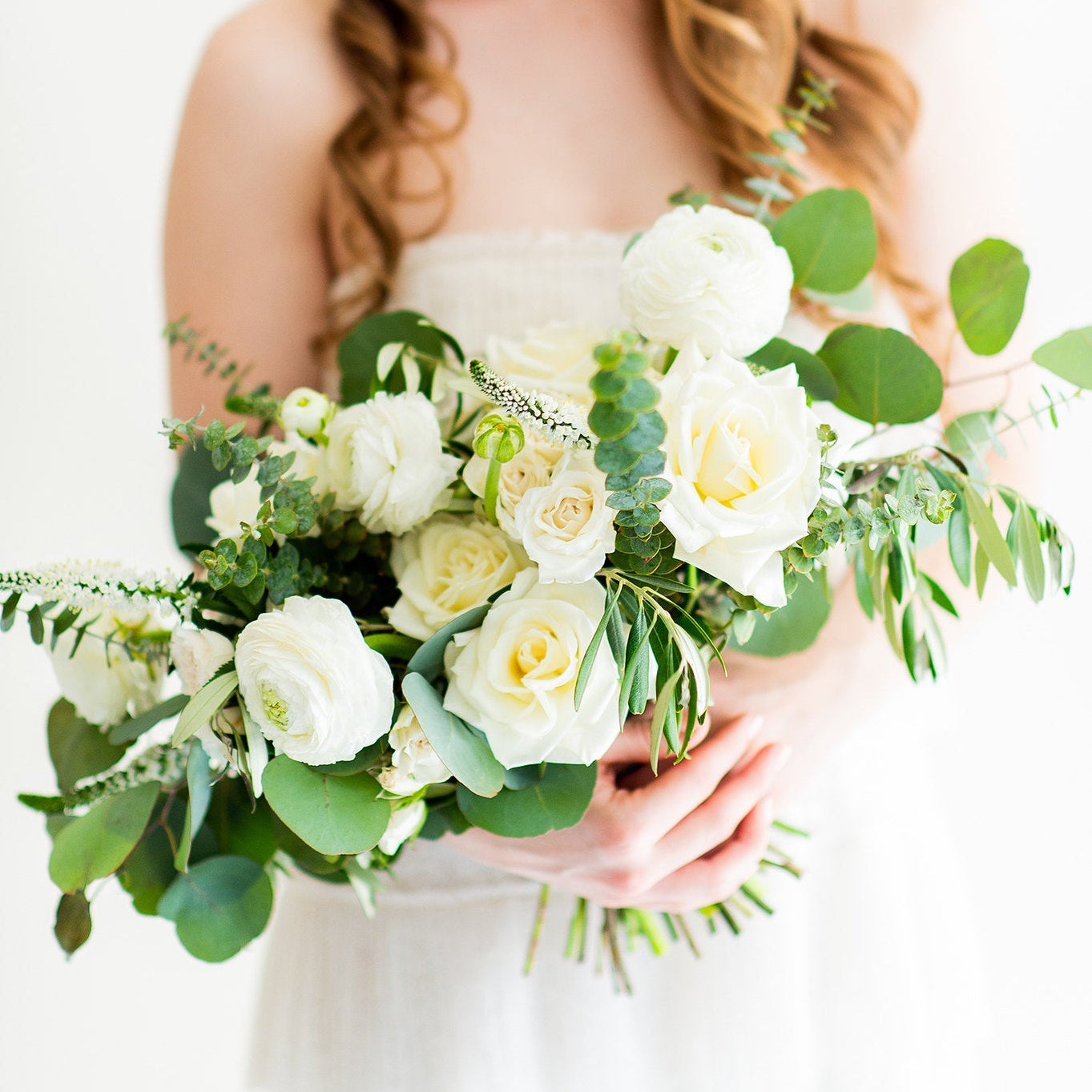 A smiling woman in a white strapless dress holds a lush bouquet of white roses and green foliage against a bright white background.