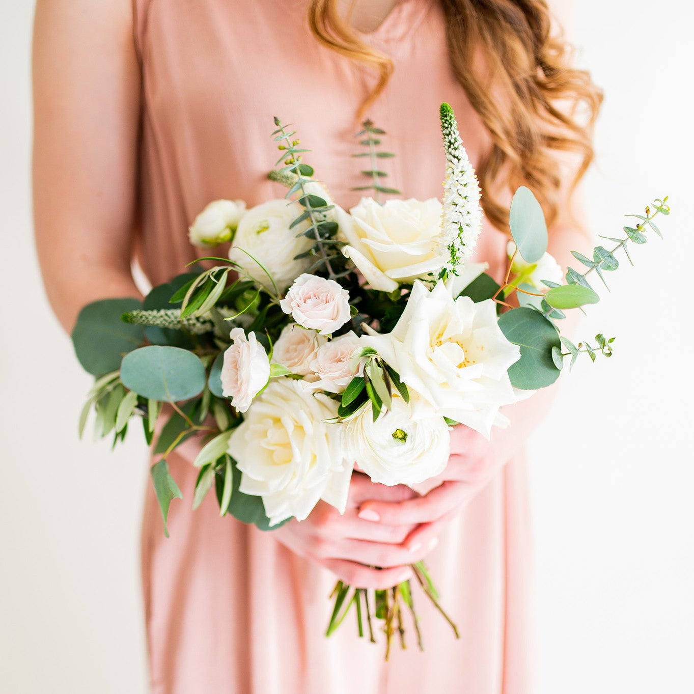 A woman in a dusty pink sleeveless dress holds a bridal bouquet of white and blush pink flowers with green foliage against a clean white background.