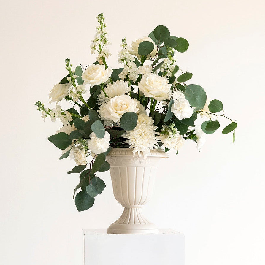A white pedestal holding a white urn-shaped vase filled with a floral arrangement of white flowers and green eucalyptus leaves, set against a plain off-white background.