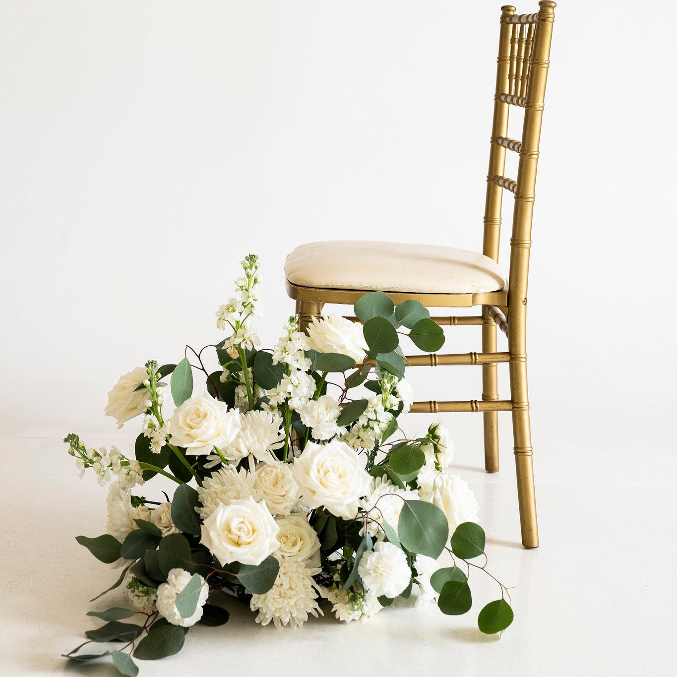 A gold-finished wooden chair with a cream upholstered seat is positioned against a seamless white studio background, with a large white floral arrangement and green foliage placed in front of it.
