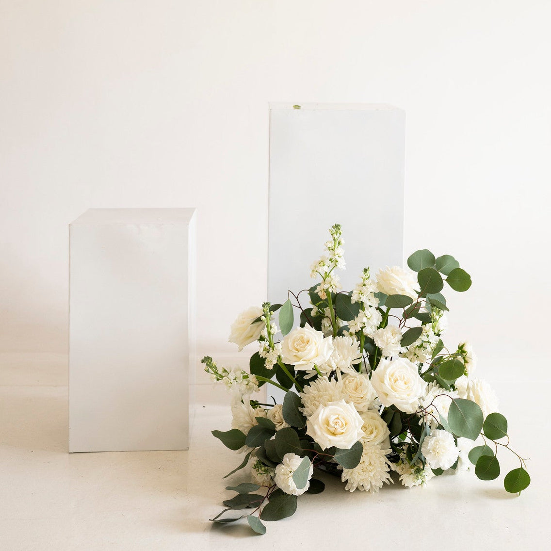 A bouquet of white flowers and green eucalyptus leaves arranged on a white surface in front of three white rectangular pedestals against a plain off-white background.