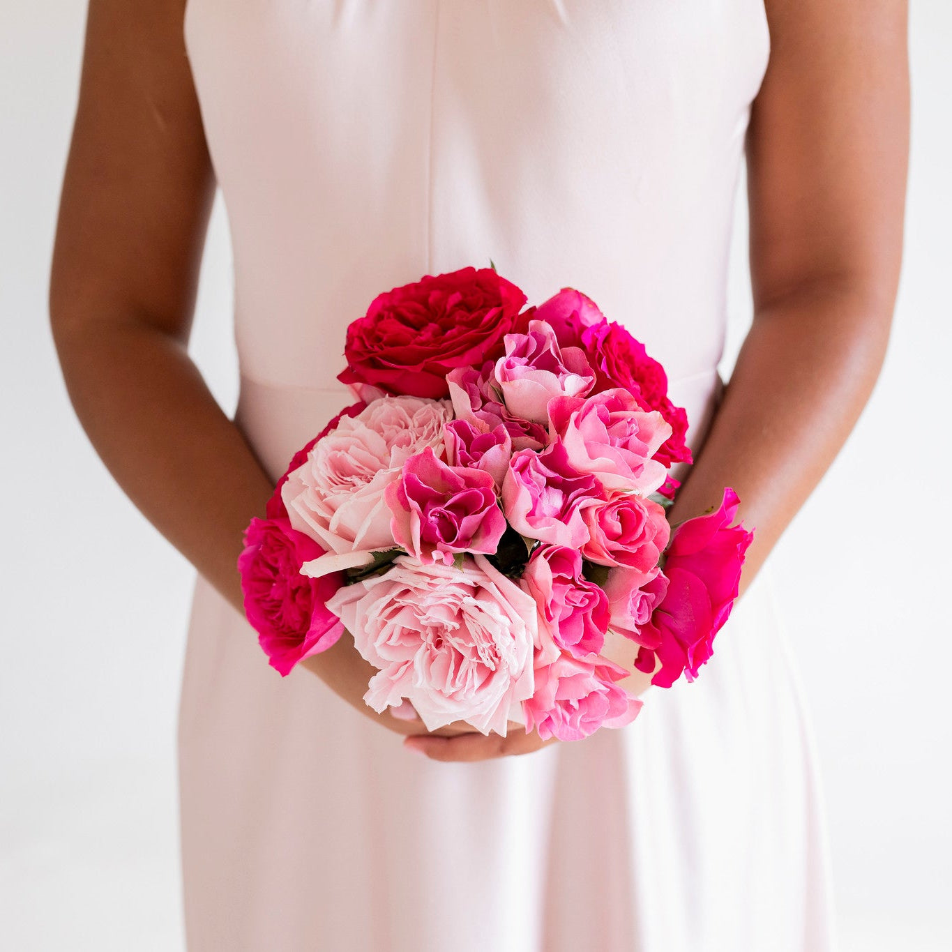 A bouquet of vibrant pink and red roses held by a person wearing a pale pink satin dress against a clean white background.