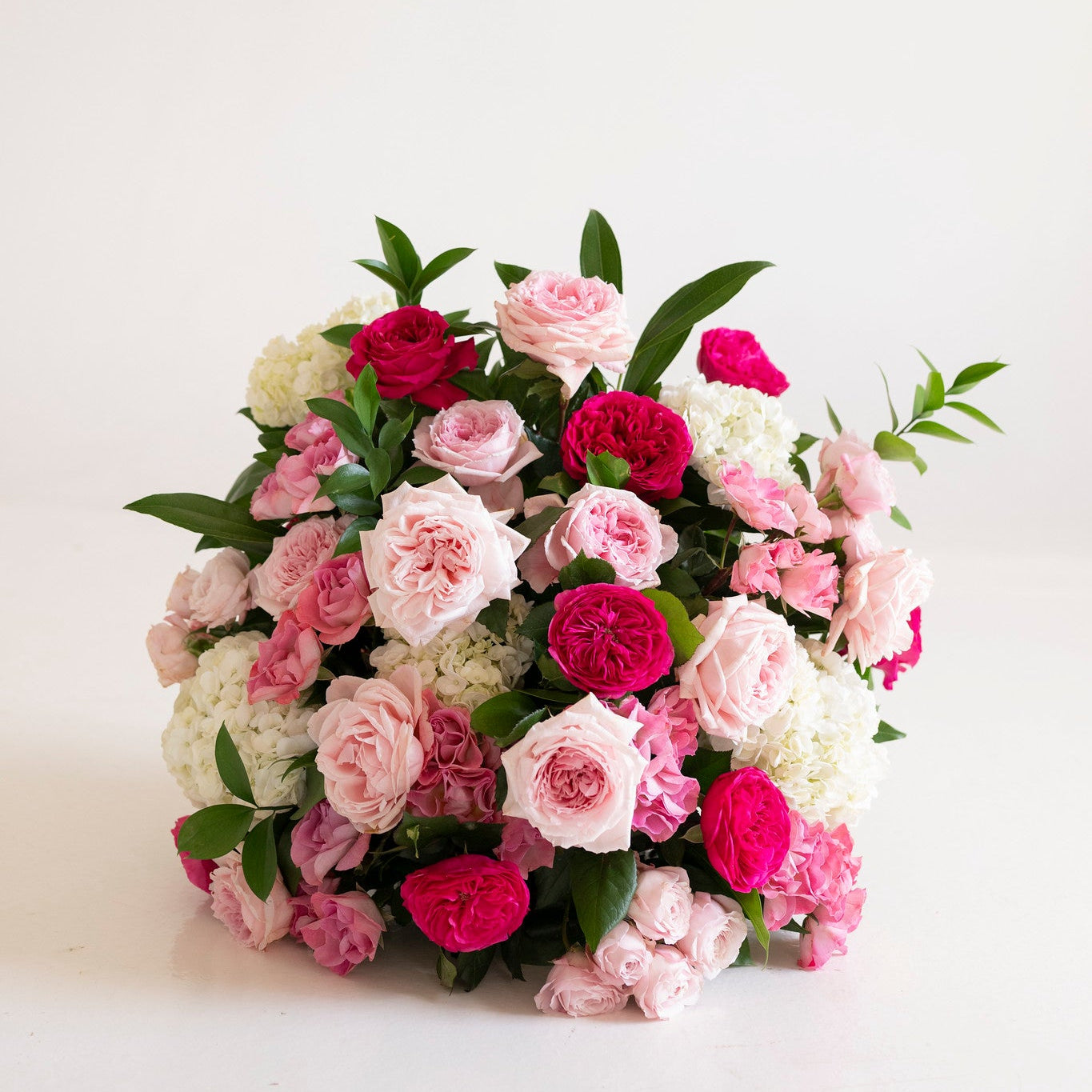 A dense, round floral arrangement featuring pink and magenta roses, white carnations, and green foliage, set against a plain white background.