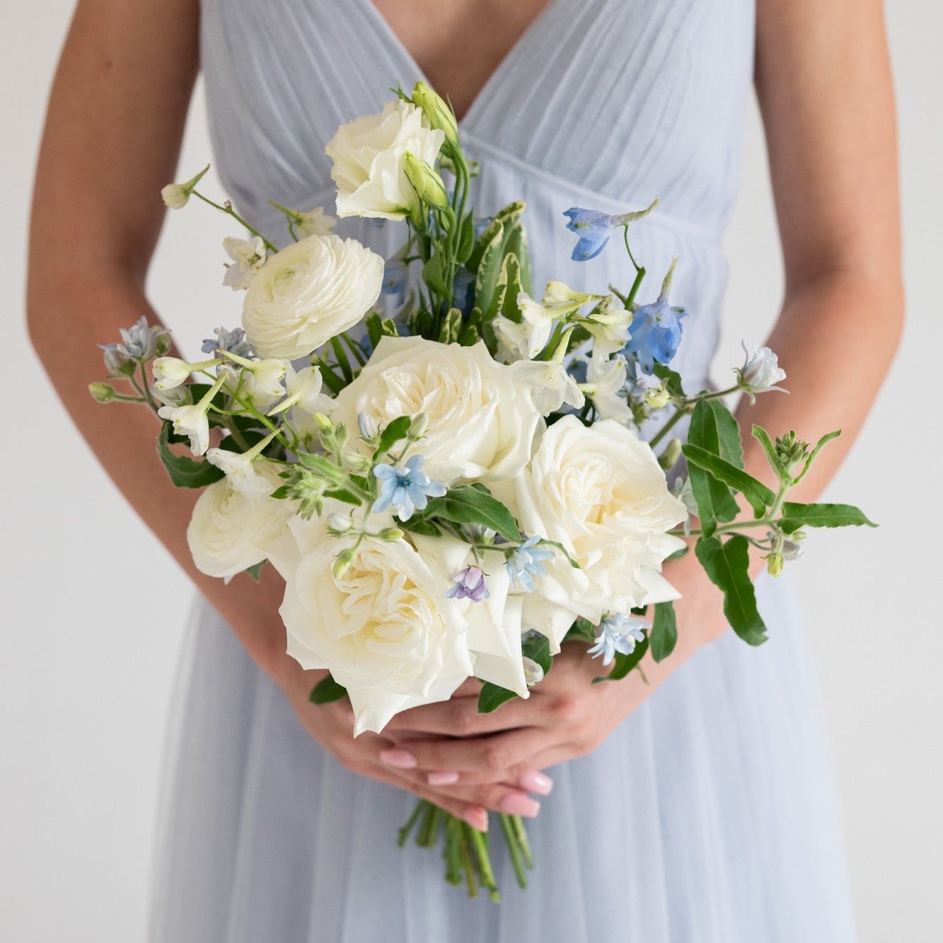 A person wearing a light blue sleeveless dress holds a bouquet of white roses and small blue flowers against a plain light background.