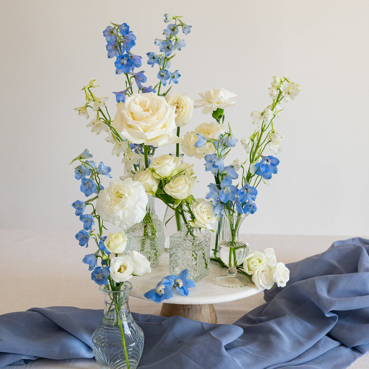 A floral arrangement of white roses, cream ranunculus, and blue delphiniums in clear glass vases, styled on a white tablecloth with a draped slate-blue fabric and a small round pedestal against a warm off-white background.