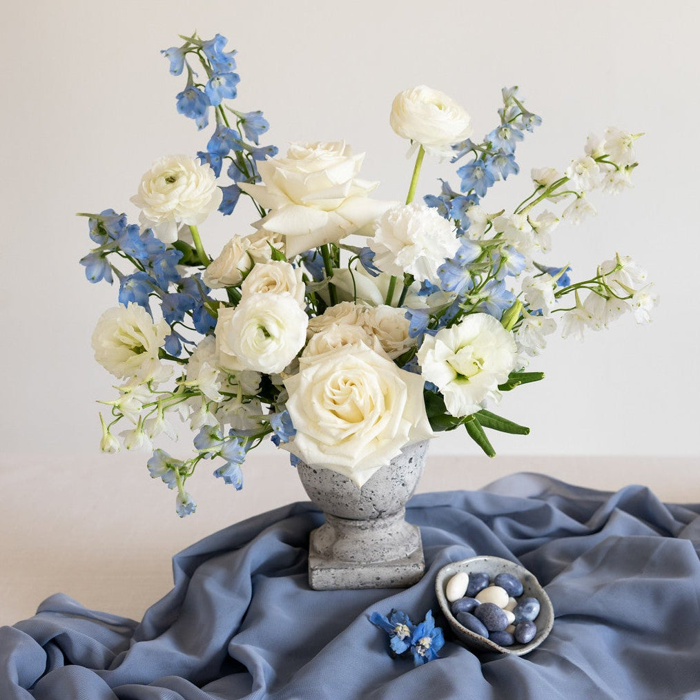 A floral arrangement of white roses and blue delphiniums in a gray stone vase, set on draped slate blue fabric with a small dish of blue flowers beside it.