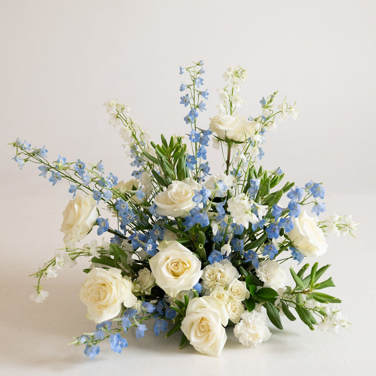 A floral arrangement featuring white roses, light blue delphiniums, and green foliage, photographed against a clean, off-white background.