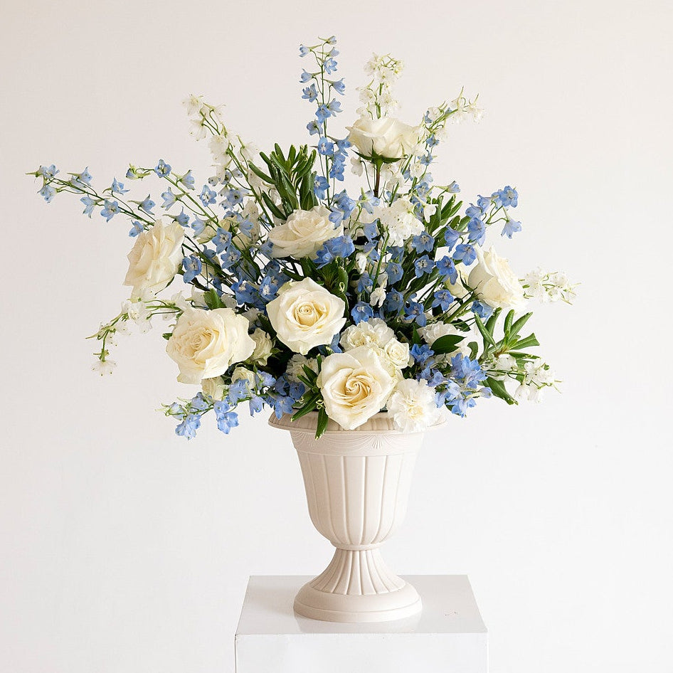 A floral arrangement featuring cream roses, blue delphiniums, and green foliage in a white classical urn vase, displayed on a white pedestal against a warm off-white background.