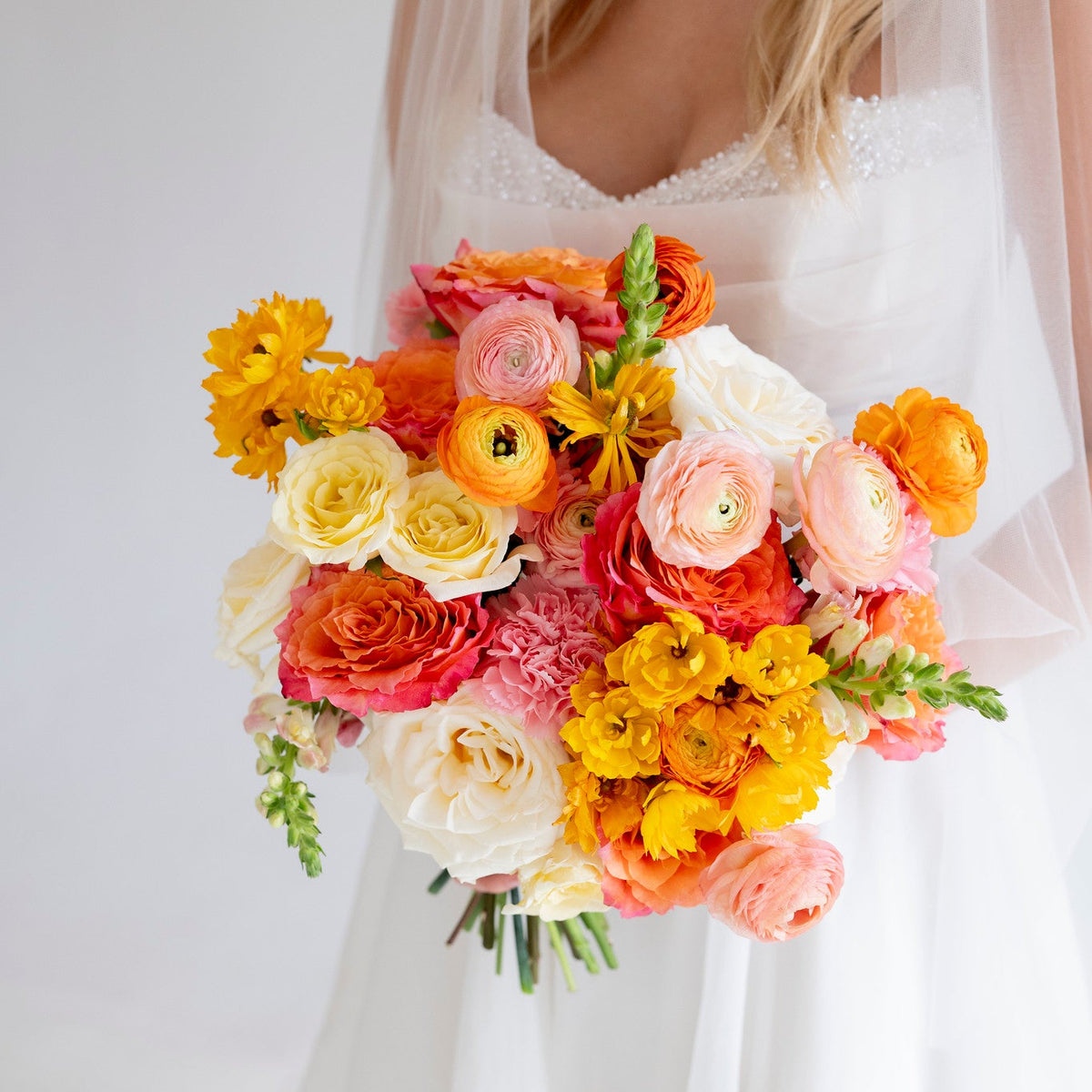 A bride in a white wedding dress and veil holds a vibrant bridal bouquet featuring orange, pink, yellow, and white flowers against a soft white background.