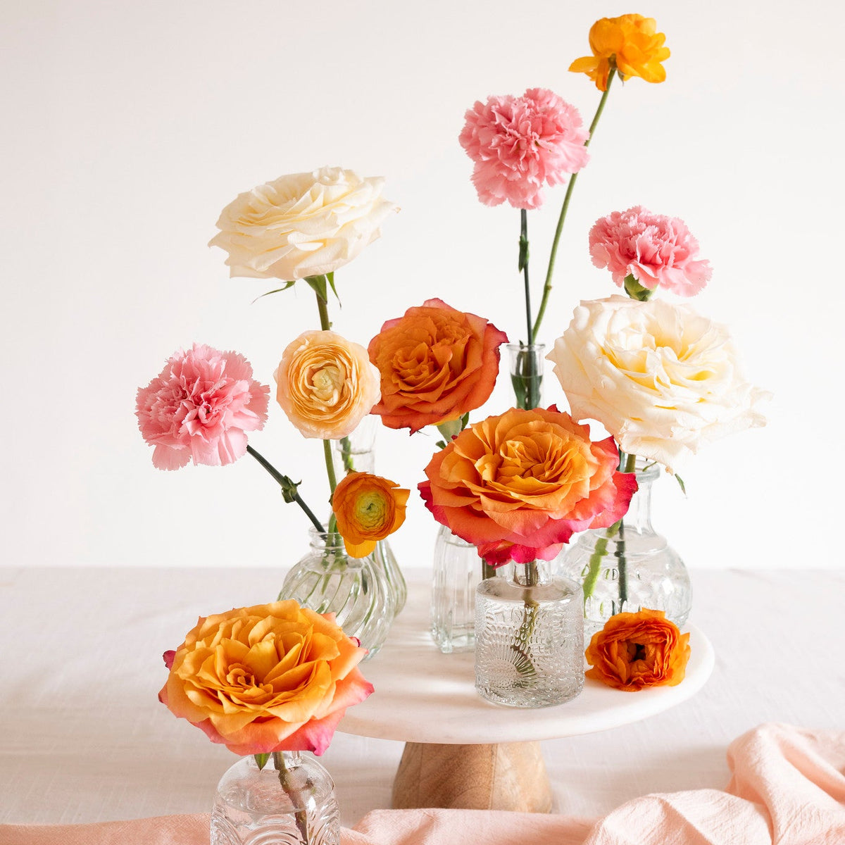 A styled floral arrangement of orange, pink, and cream flowers in clear glass bottles and on a white pedestal stand, set against a clean white background with a draped peach fabric.