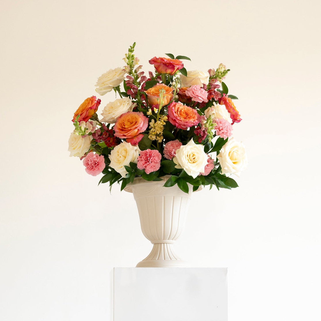 A vibrant artificial flower bouquet in a white classical-style urn vase, displayed on a white pedestal against a clean white background.