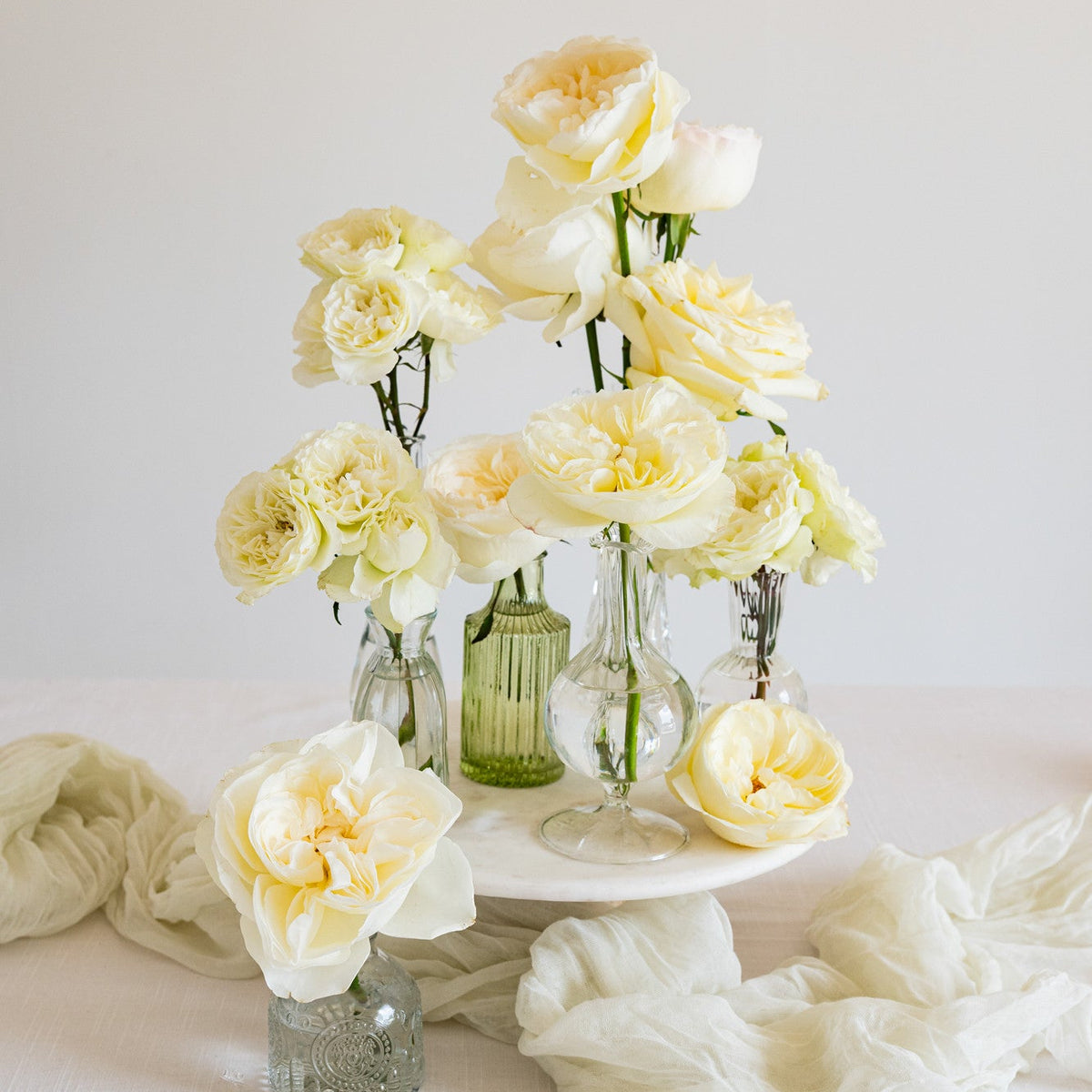 A styled floral arrangement of pale cream roses and carnations in clear glass vases and a small green bottle, displayed on a white pedestal with draped white fabric against a soft off-white background.