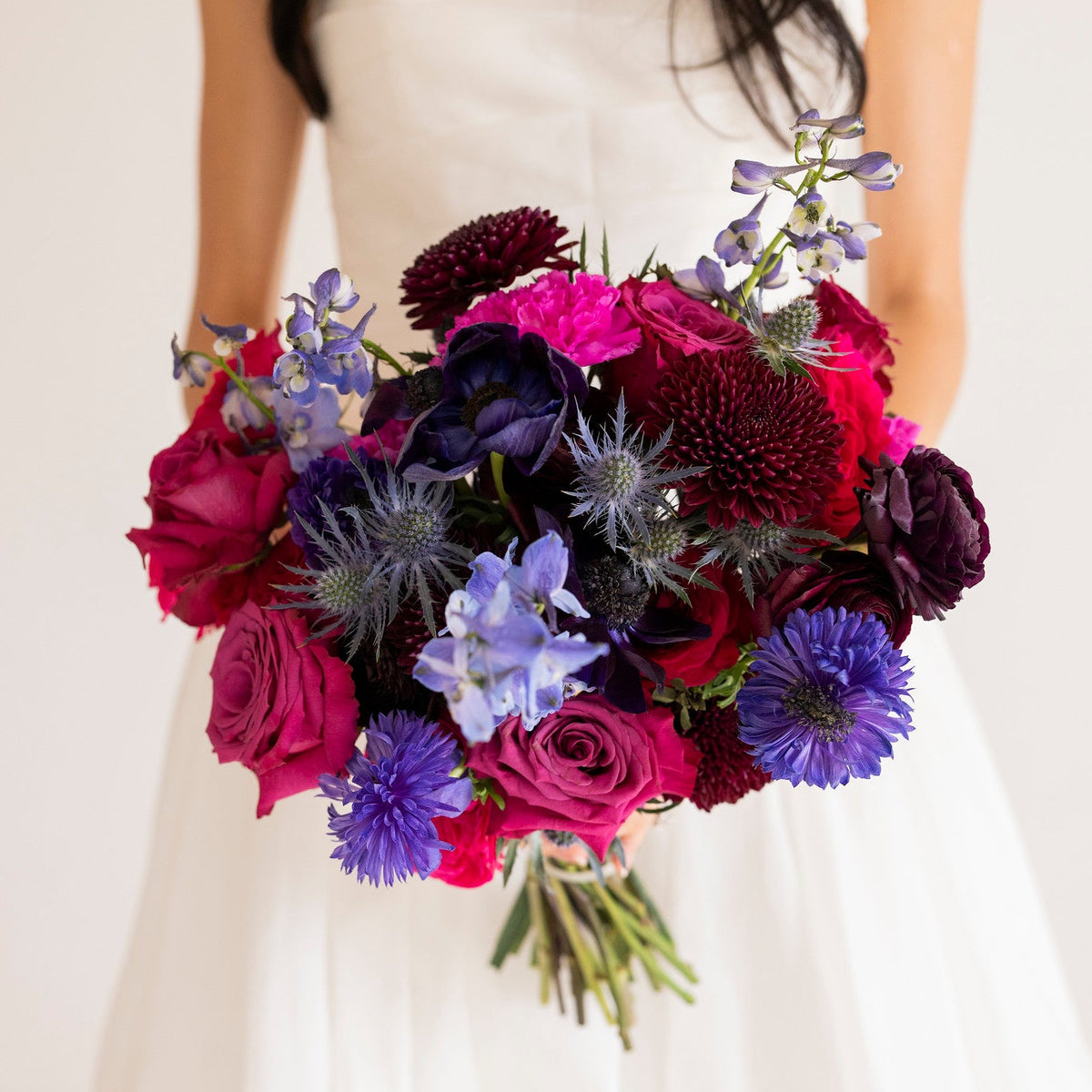 A close-up of a woman in a white strapless wedding dress holding a vibrant bouquet of deep magenta, purple, and blue flowers against a soft off-white background.