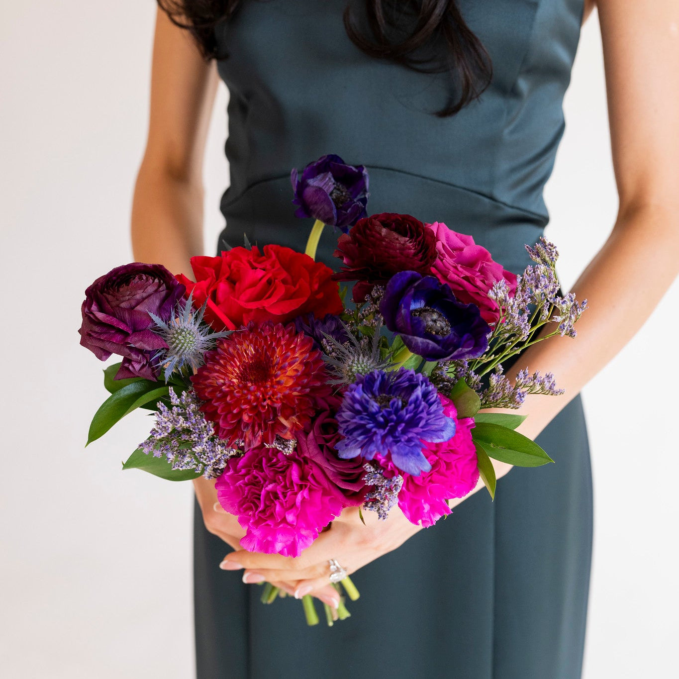 A woman in a dark teal sleeveless dress holds a vibrant, multicolored floral bouquet featuring bright pink, deep red, purple, and blue flowers with green leaves, set against a plain off-white background.