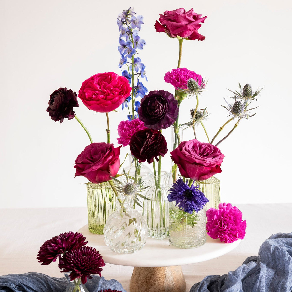 A floral arrangement of pink, purple, burgundy, and blue flowers in clear glass vases, displayed on a white cake stand with a blue-gray draped fabric in the foreground.