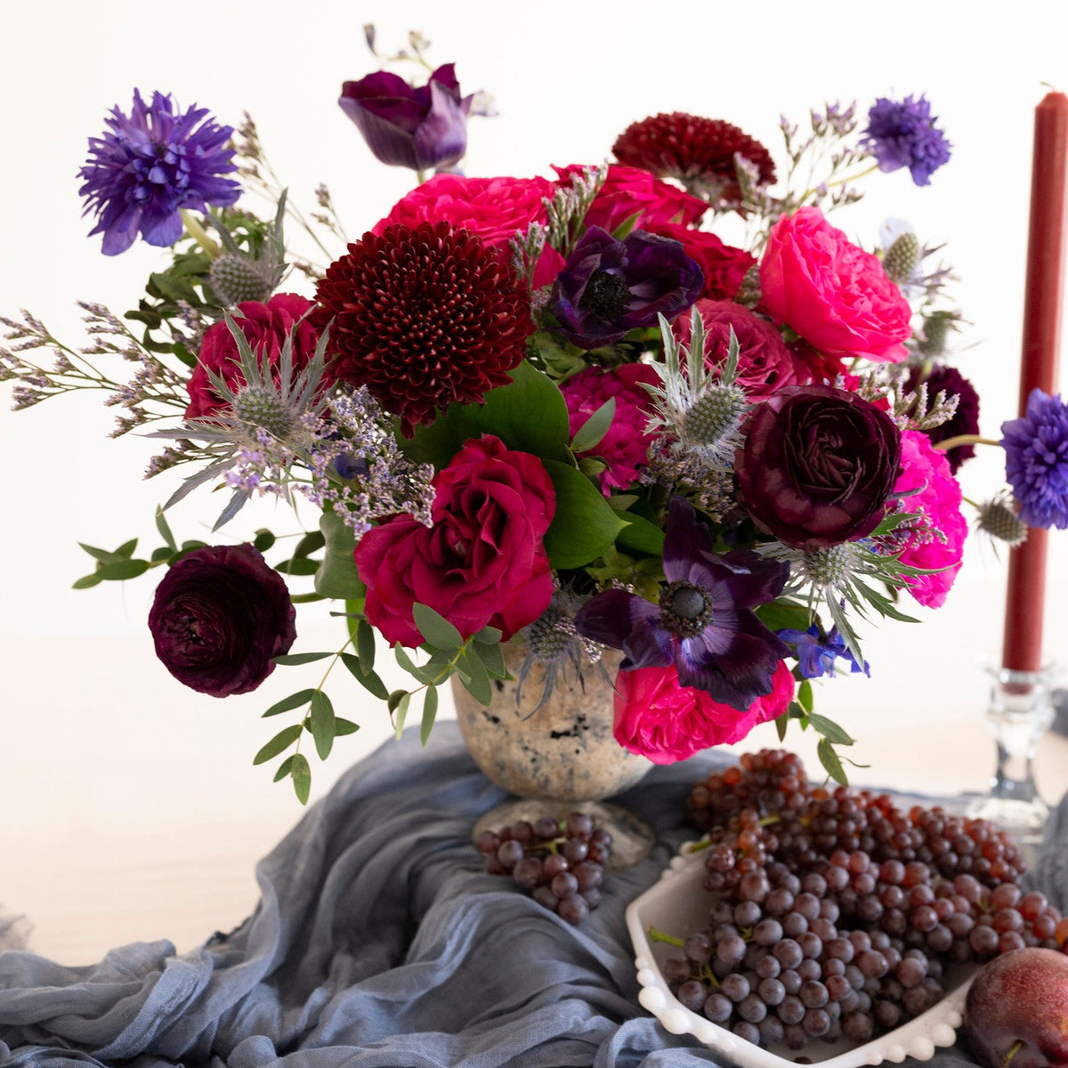 A vibrant floral arrangement featuring deep magenta, burgundy, and purple flowers with green foliage, presented in a textured vase draped over a slate blue fabric, with grapes and a red candle as styling props.