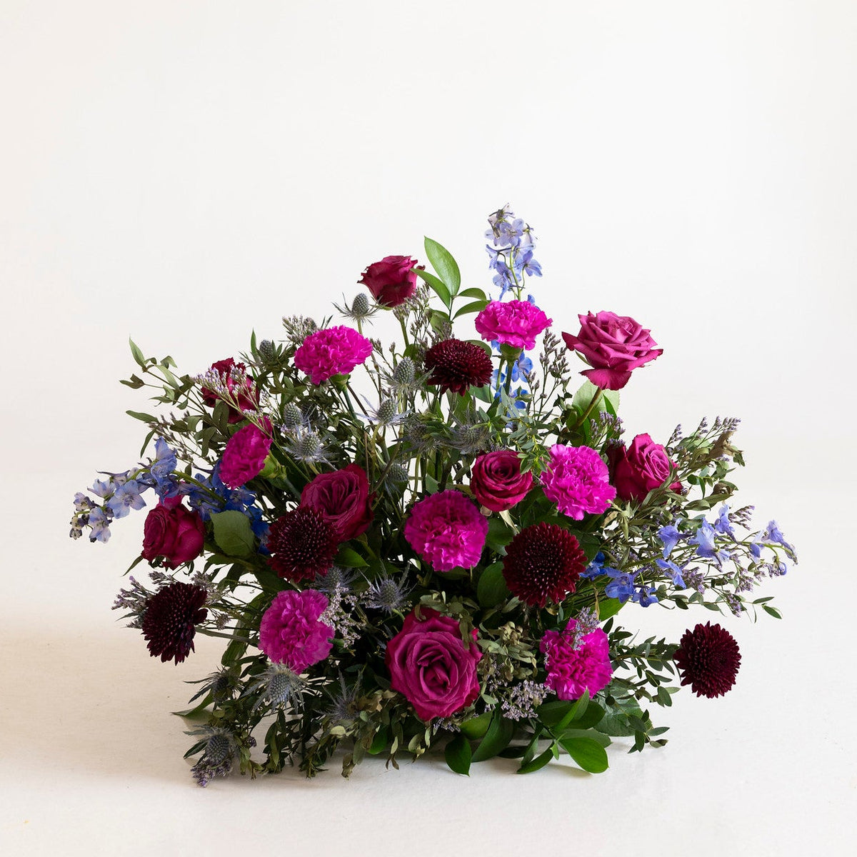 A vibrant floral arrangement featuring magenta and deep burgundy roses, blue delphiniums, and green foliage, set against a clean off-white background.