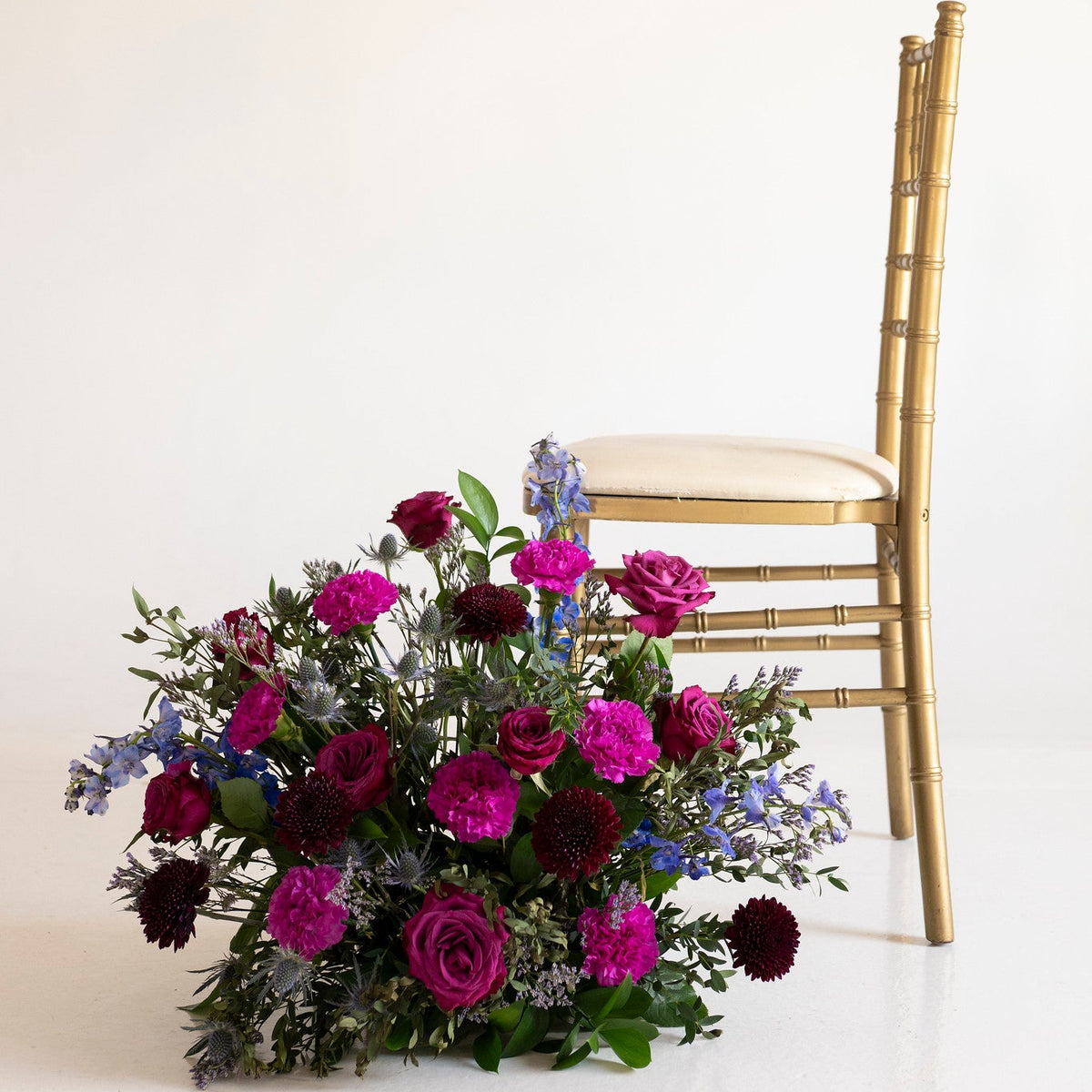 A large floral arrangement of magenta, burgundy, and blue flowers with green foliage, placed on the floor beside a gold Chiavari chair against a plain off-white background.