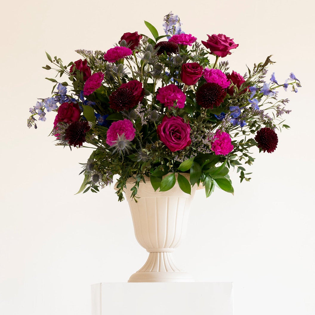 A vibrant floral arrangement featuring magenta and burgundy roses with green foliage, presented in a white classical-style urn on a white pedestal against a clean white background.
