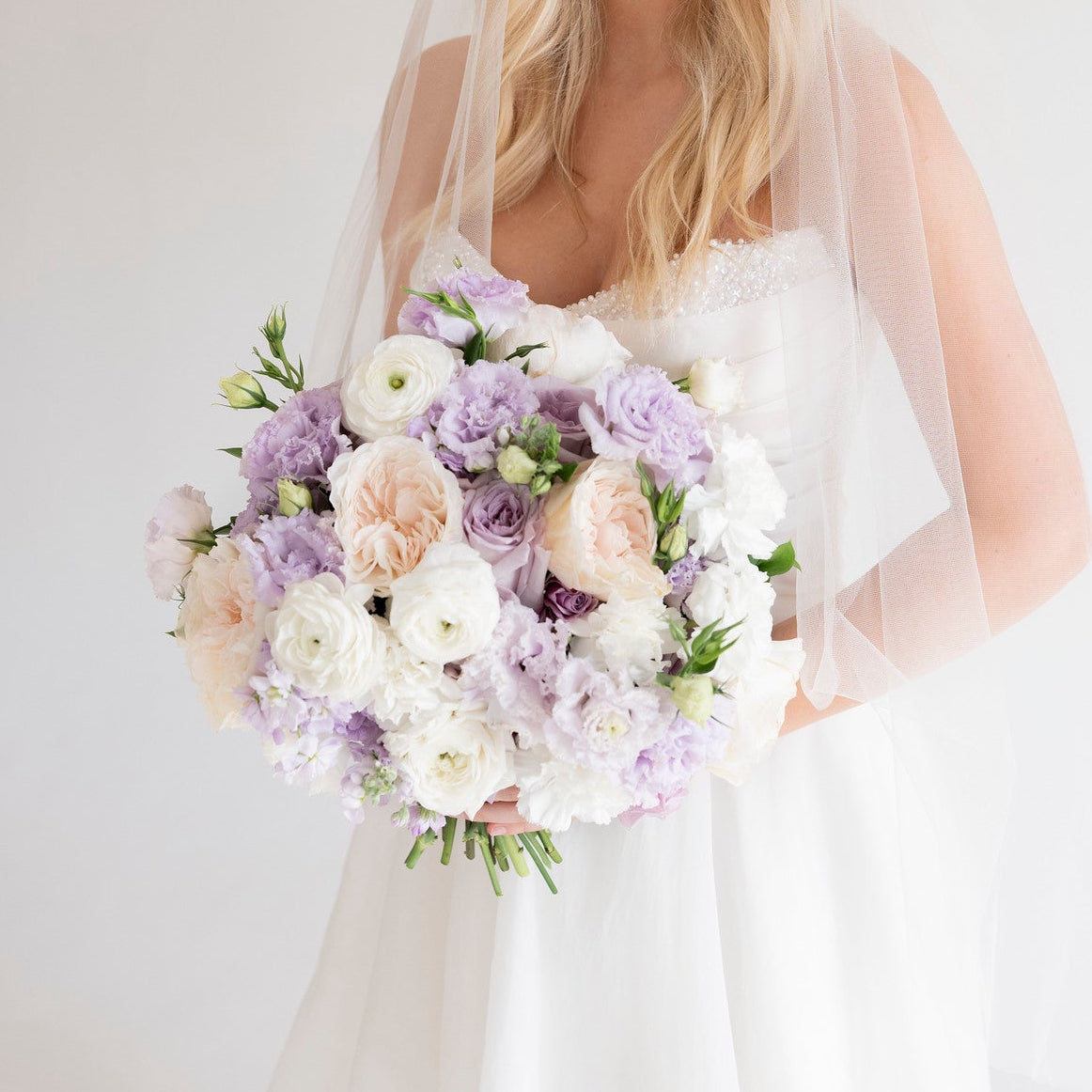 A smiling blonde bride in a white wedding dress and veil holds a large bouquet of white, lavender, and blush flowers against a soft light gray studio background.