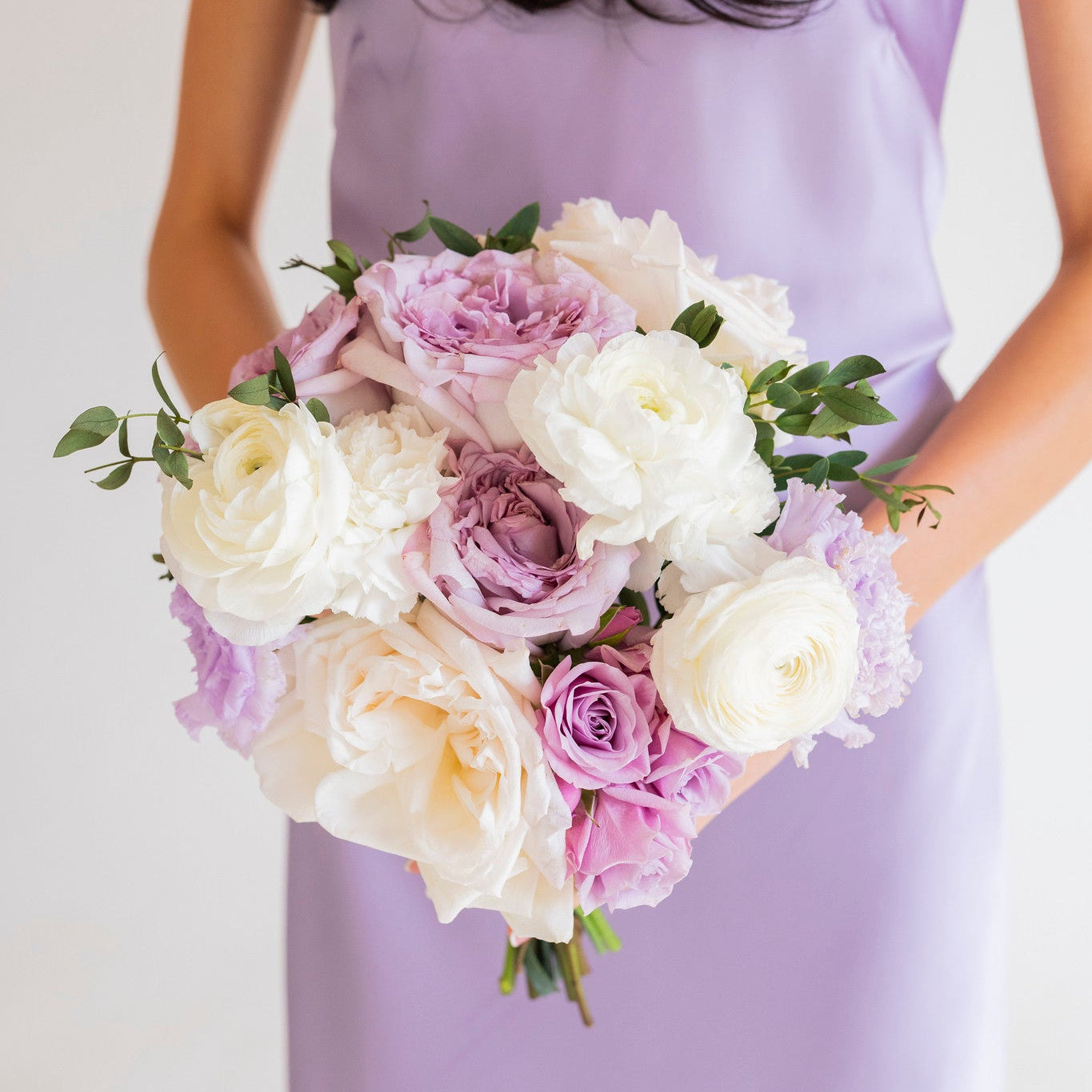 A woman in a lavender sleeveless dress holds a bouquet of white and lavender roses with green leaves against a plain light background.