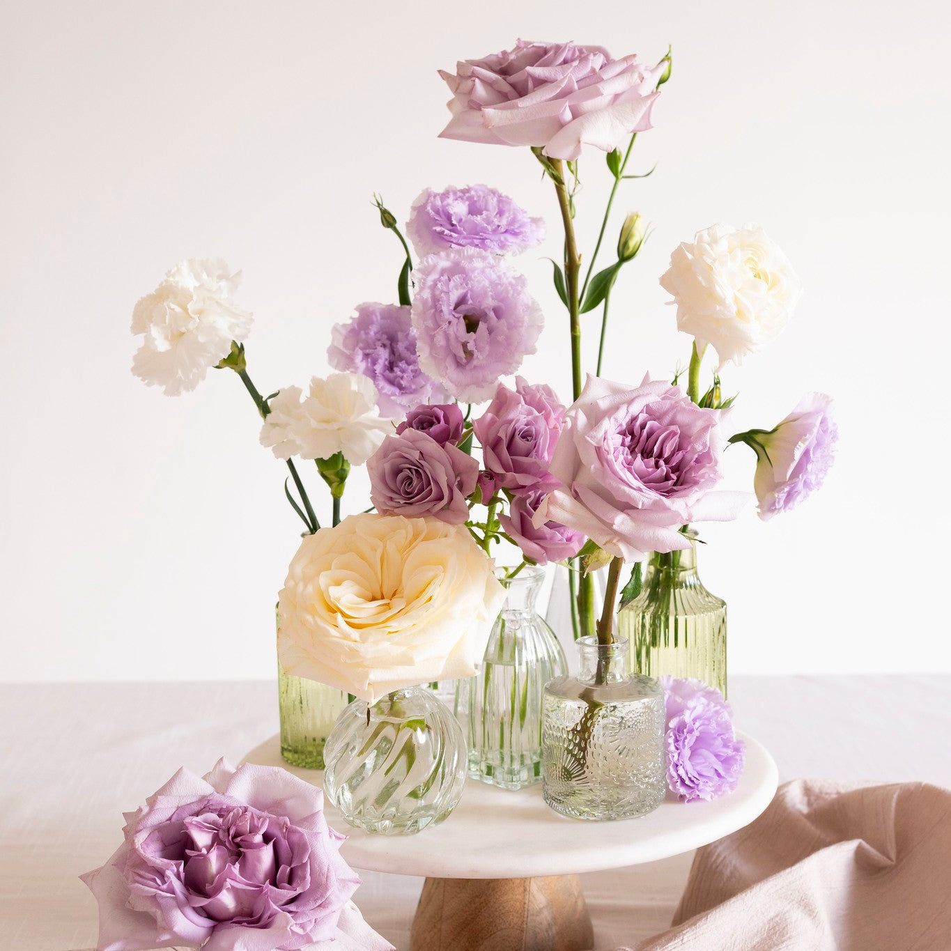 A styled floral arrangement of lavender, lilac, cream, and white flowers in clear glass bottles and a small vase, displayed on a white pedestal stand with a draped blush pink fabric against a soft off-white background.