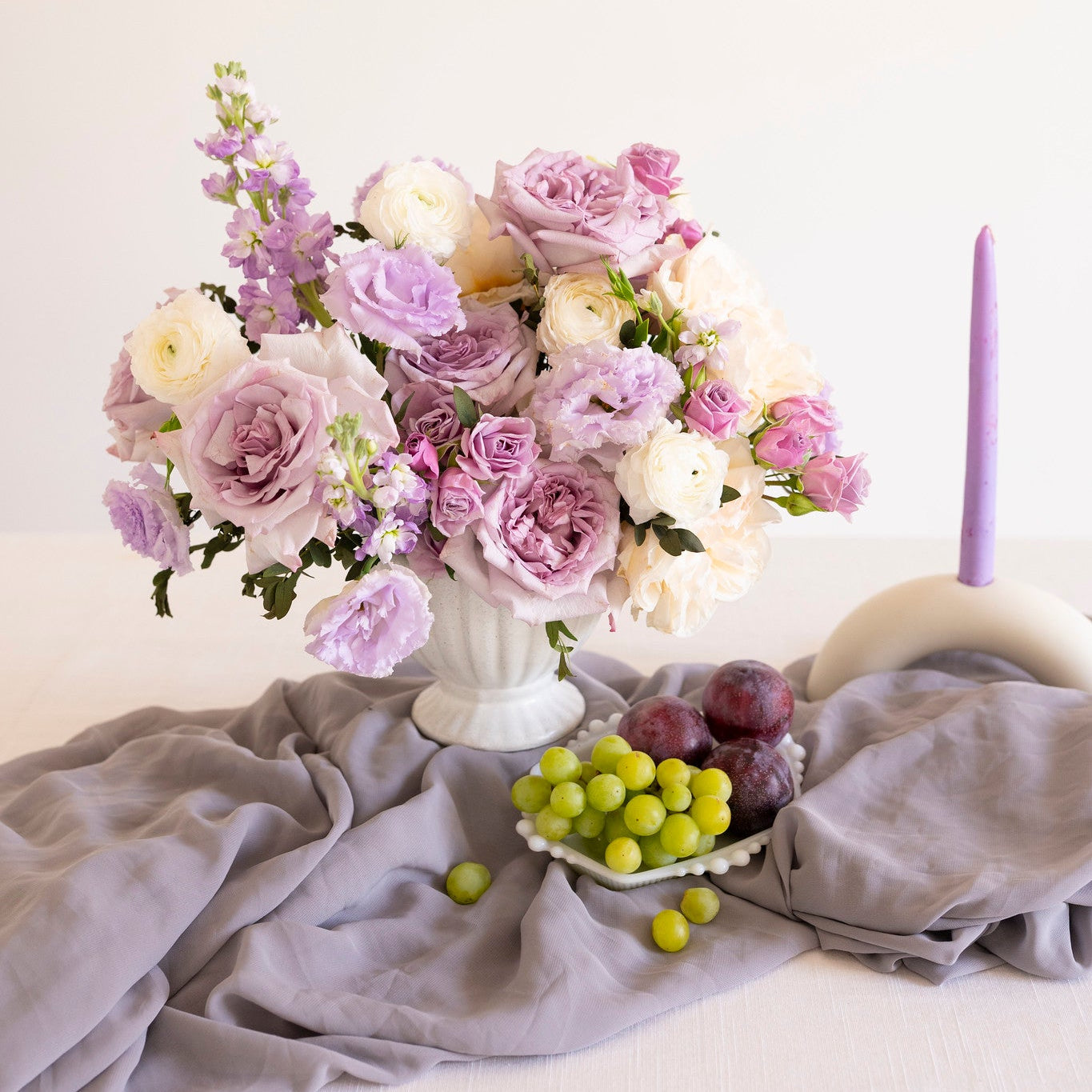 A styled floral arrangement in a white vase with lavender, blush, and cream flowers, set on a draped gray fabric surface with green grapes and a lit purple taper candle in a ceramic holder against a plain light background.