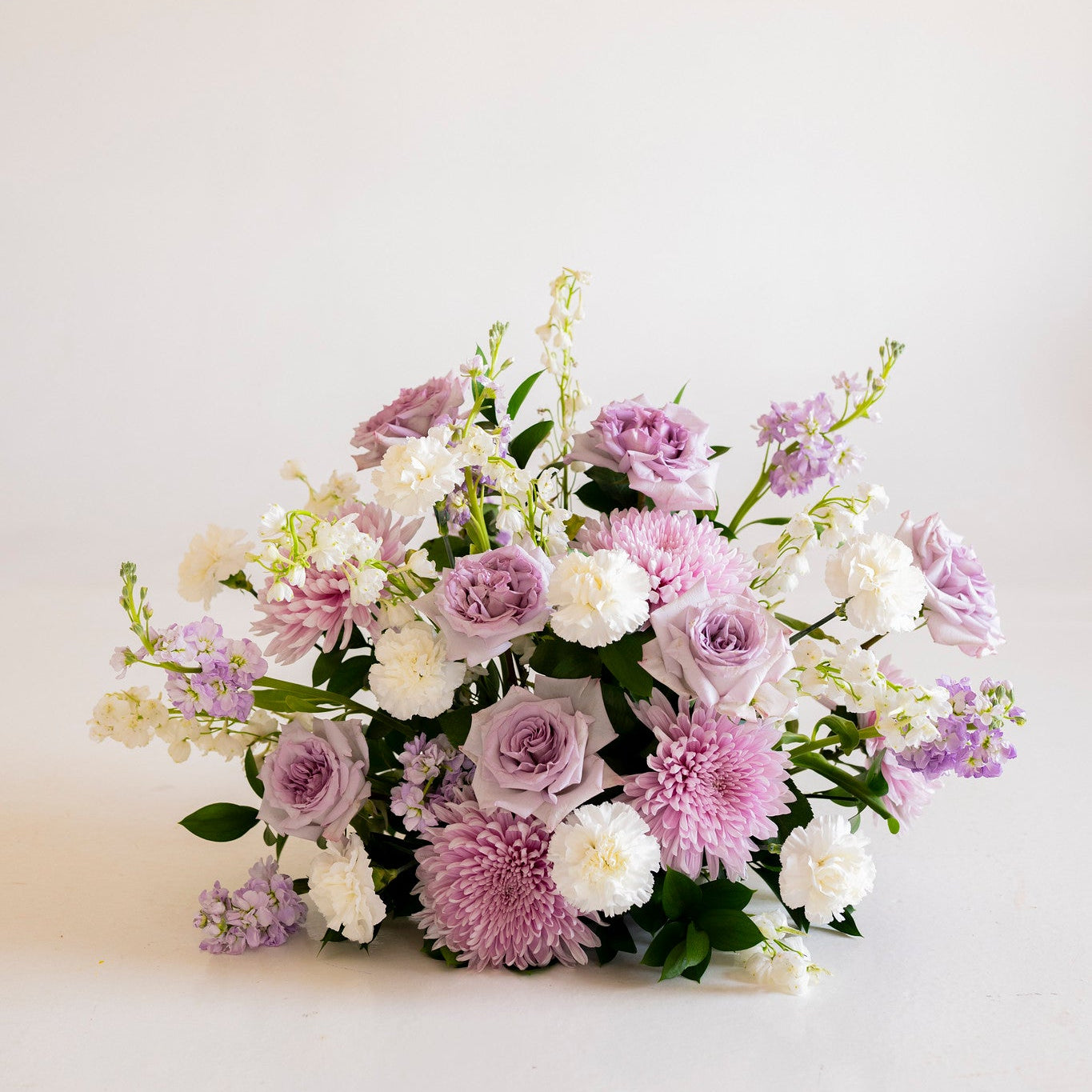 A mixed floral arrangement featuring light purple, white, and pink flowers with green foliage, photographed on a clean white studio background.