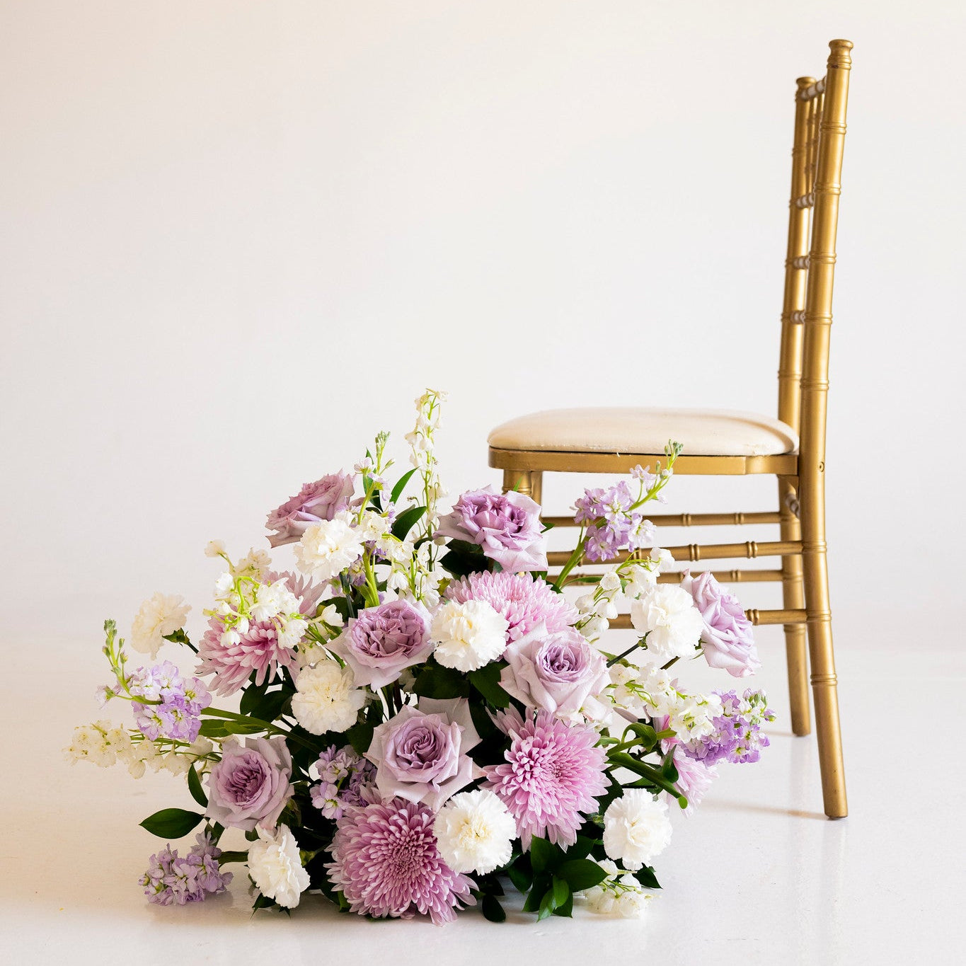 A floral arrangement of lavender, white, and purple flowers with green foliage placed on a white surface beside a gold chair against a plain light background.