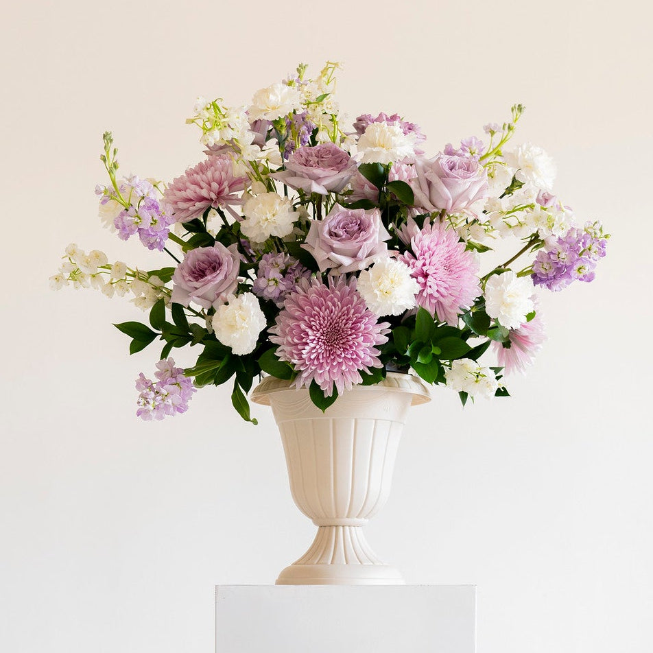 A floral arrangement of pink, purple, and white flowers with green foliage, displayed in a white urn-shaped vase on a white pedestal against a plain light background.