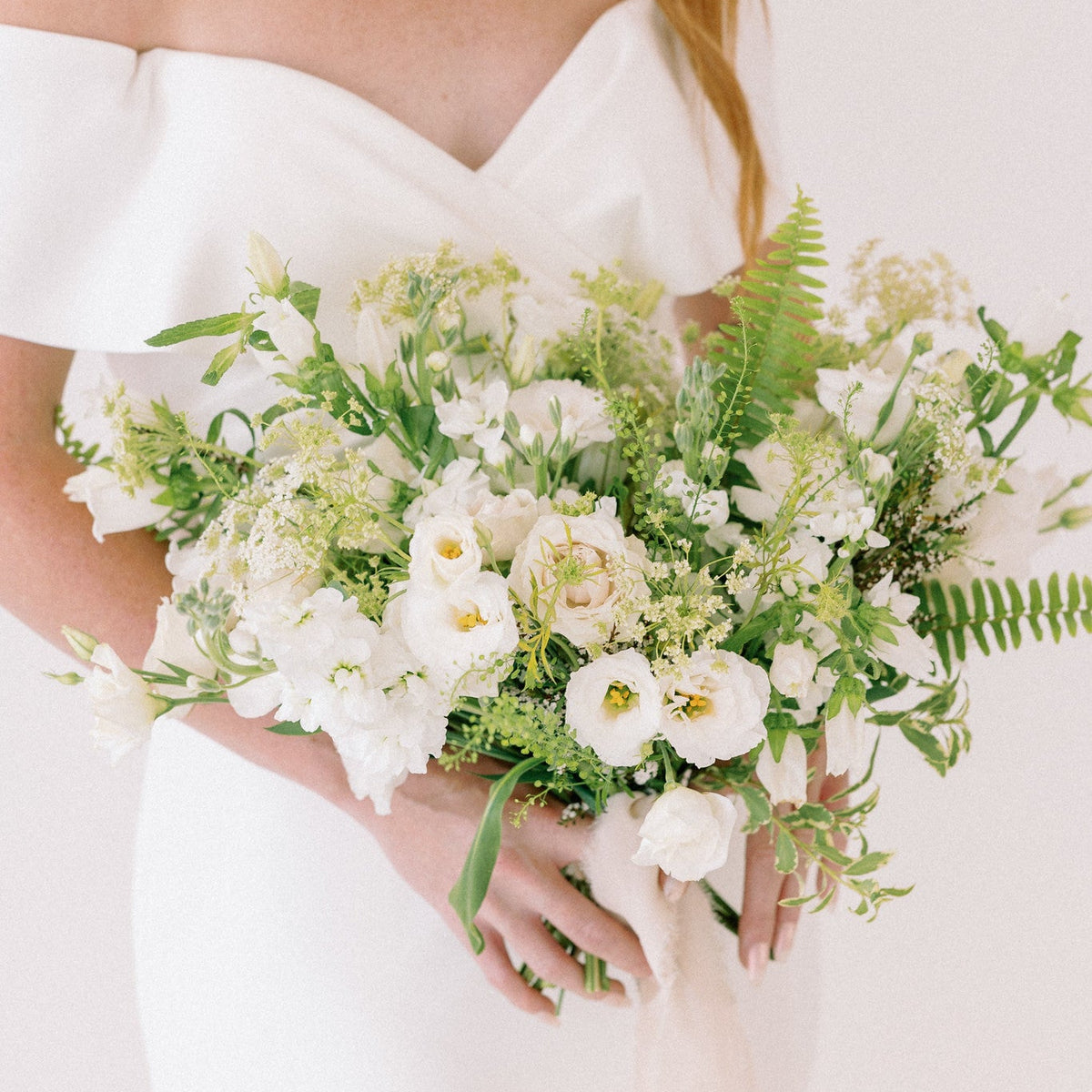 A woman in a white off-the-shoulder dress holding a large bridal bouquet of white flowers and green foliage against a plain light background.