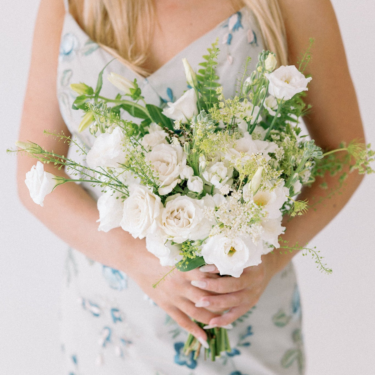 A woman holding a large bouquet of white flowers and green foliage, wearing a sleeveless white dress with a blue floral pattern against a plain light background.