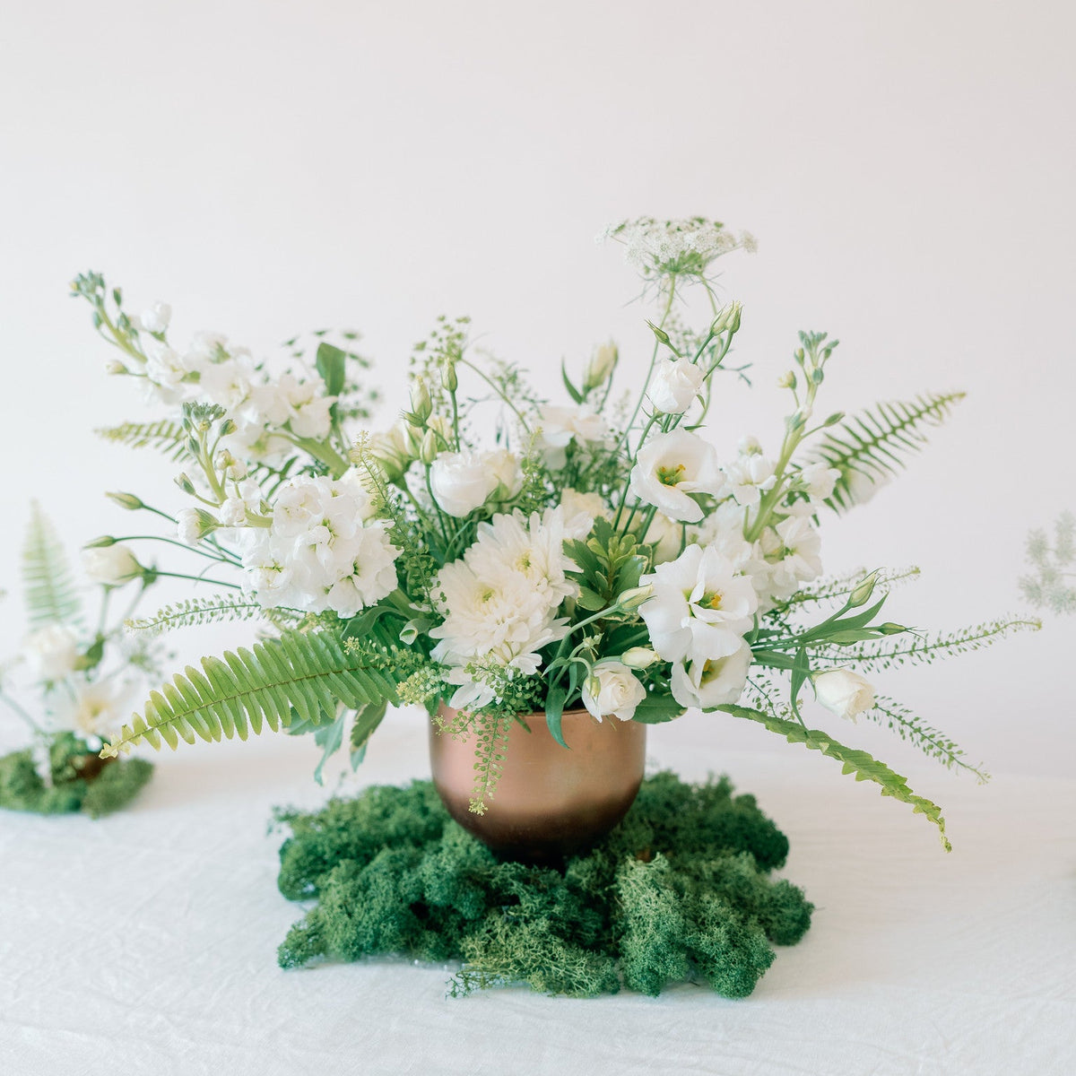 A floral centerpiece arrangement with white flowers and green foliage in a metallic bronze pot set on a bed of green moss, photographed on a white tablecloth against a soft white background.