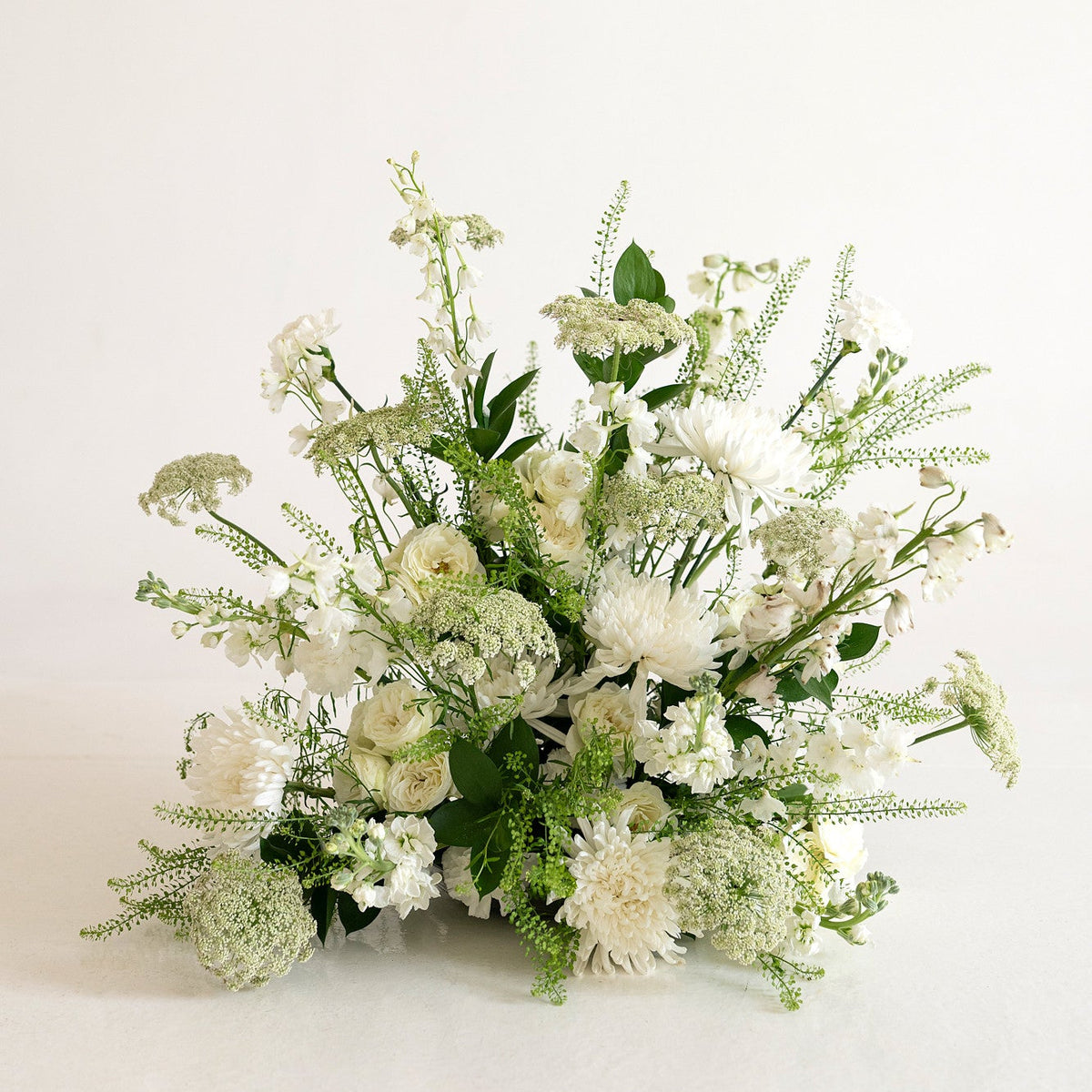 A white and green floral arrangement featuring white flowers and lush green foliage, photographed against a seamless white studio background.