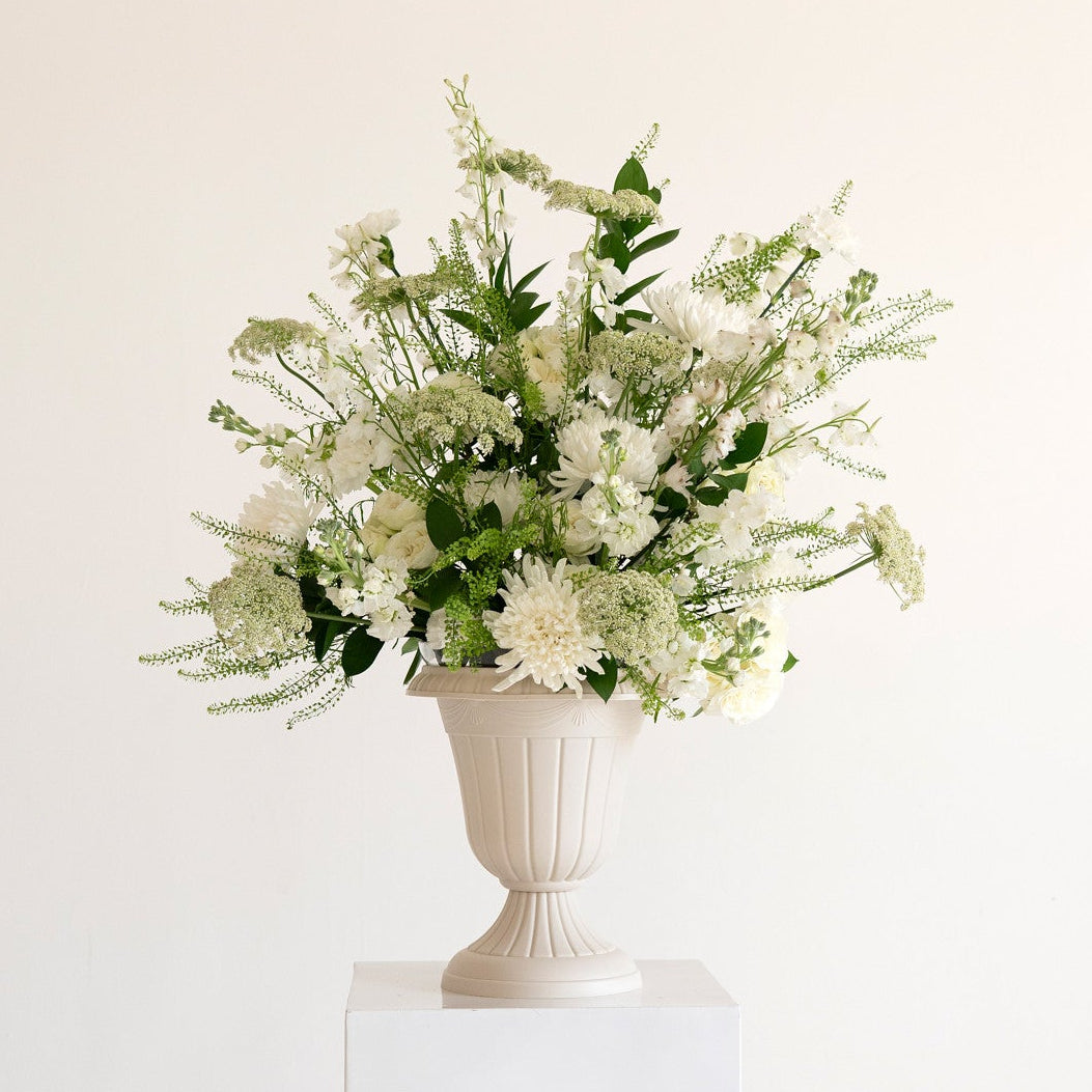 A white ceramic urn-shaped vase filled with a lush bouquet of white flowers and green foliage, displayed on a white pedestal against a clean white background.