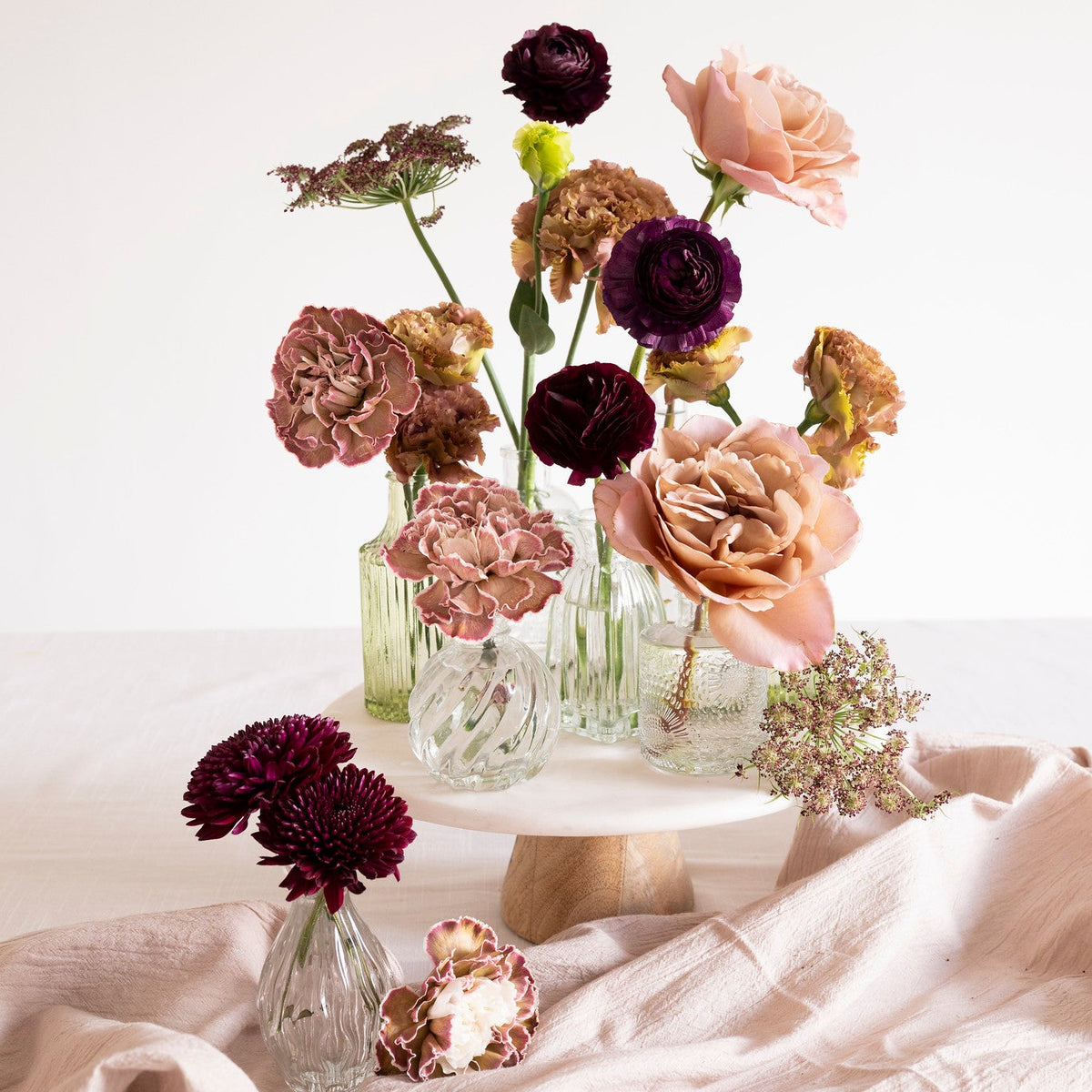 A styled floral arrangement of dried and fresh flowers in clear glass vases on a white pedestal stand, with additional flowers and a small vase placed on draped blush pink fabric against a clean white background.