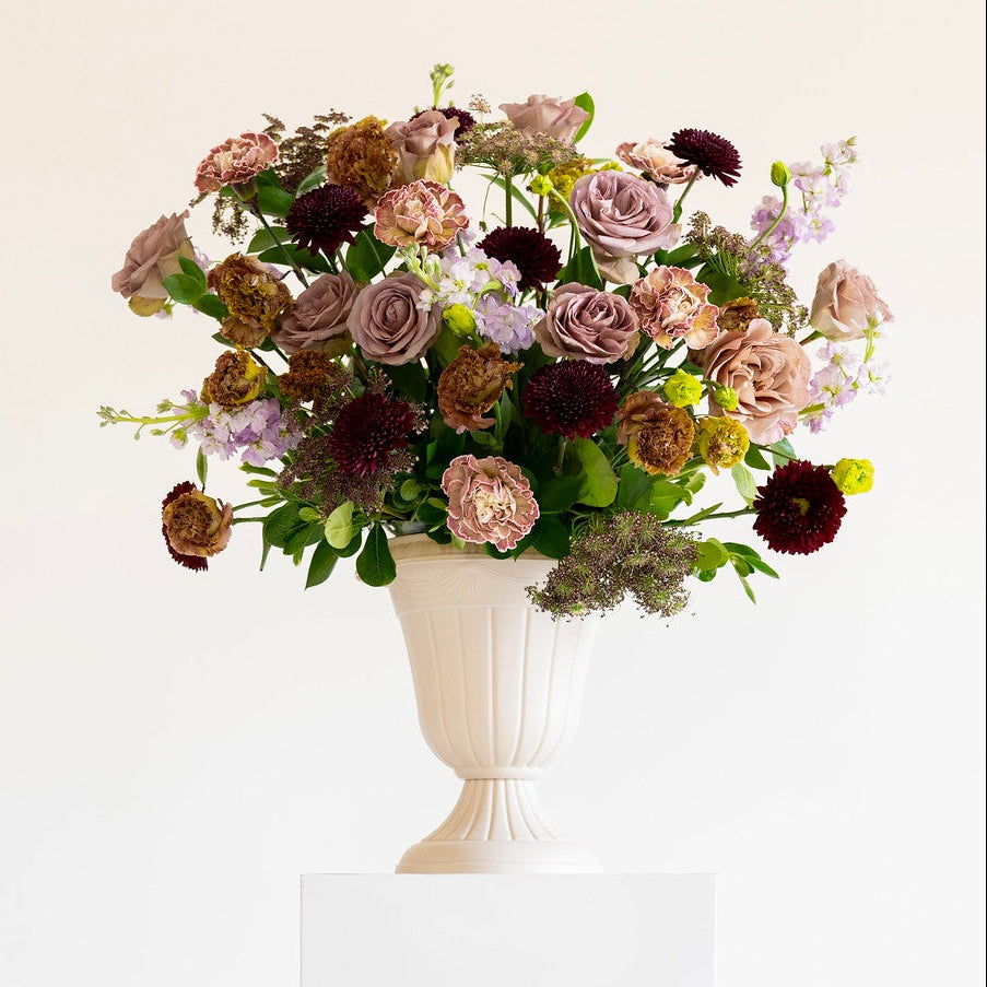 A floral arrangement in a white pedestal vase, featuring muted pink, burgundy, and purple flowers with green foliage, displayed on a white rectangular plinth against a clean white background.