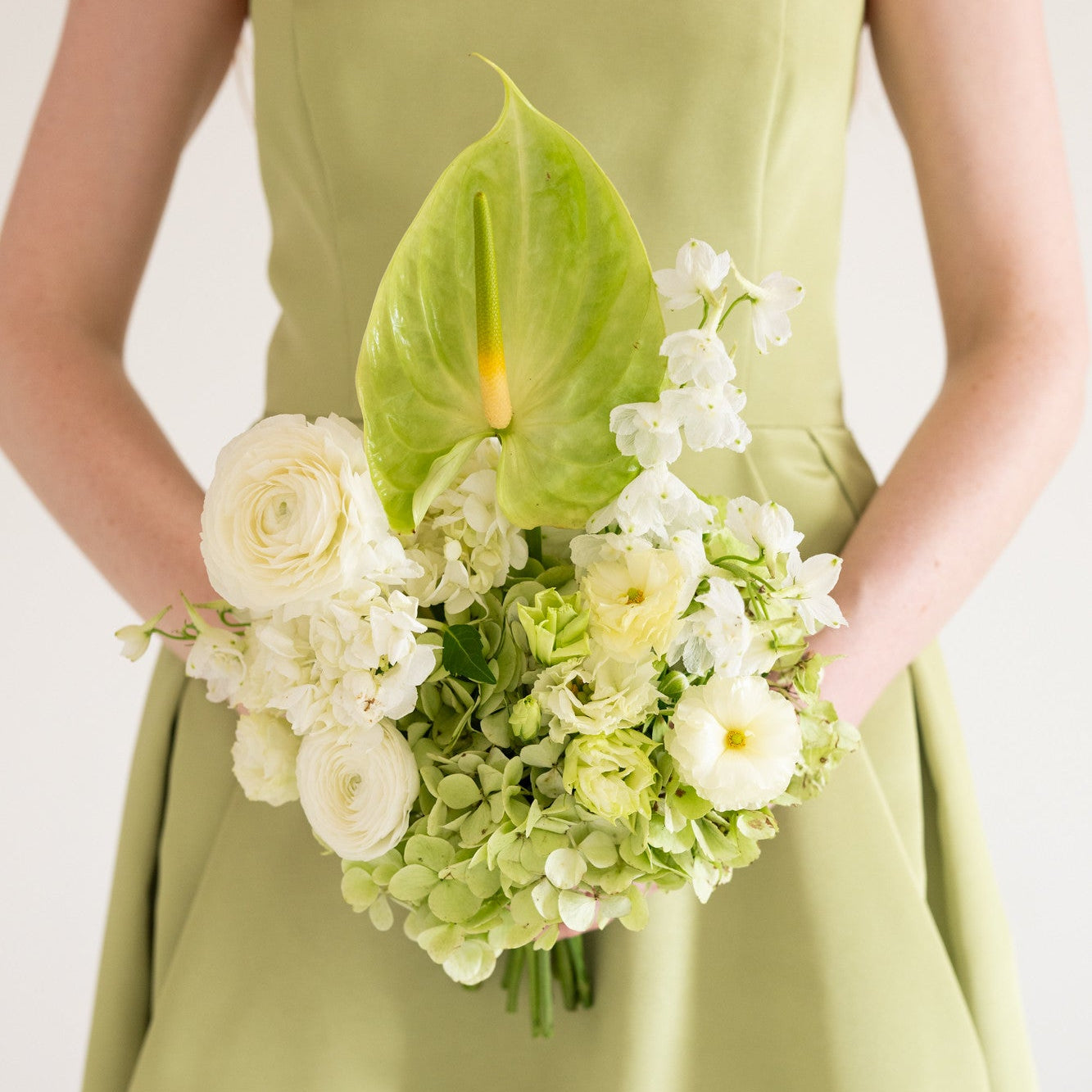 A person wearing a light olive green dress holds a bouquet of white and pale green flowers, including a large green leaf and white roses, against a plain light background.