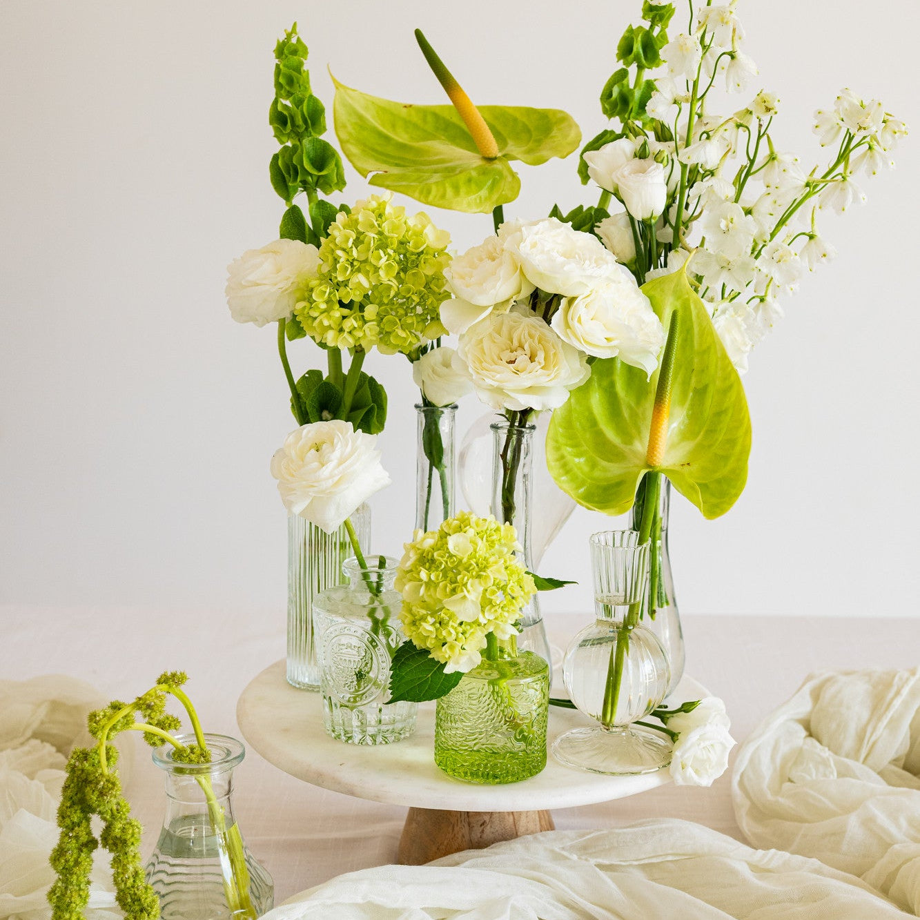 A floral arrangement of white and green flowers in clear glass vases and a small green jar, displayed on a round white pedestal stand with draped white fabric on a light surface against a soft white background.