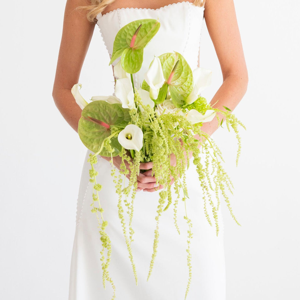 A smiling blonde woman in a white strapless dress holds a lush green and white floral bouquet with trailing vines against a clean white studio background.