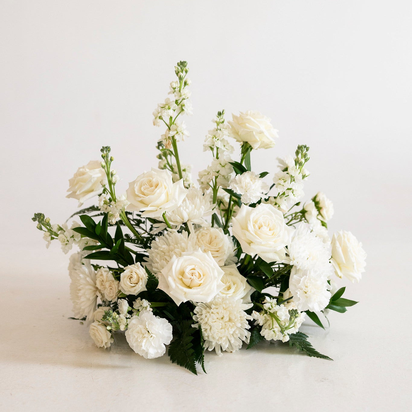 A white floral arrangement featuring roses, chrysanthemums, and delphiniums with green foliage, photographed against a clean white background.
