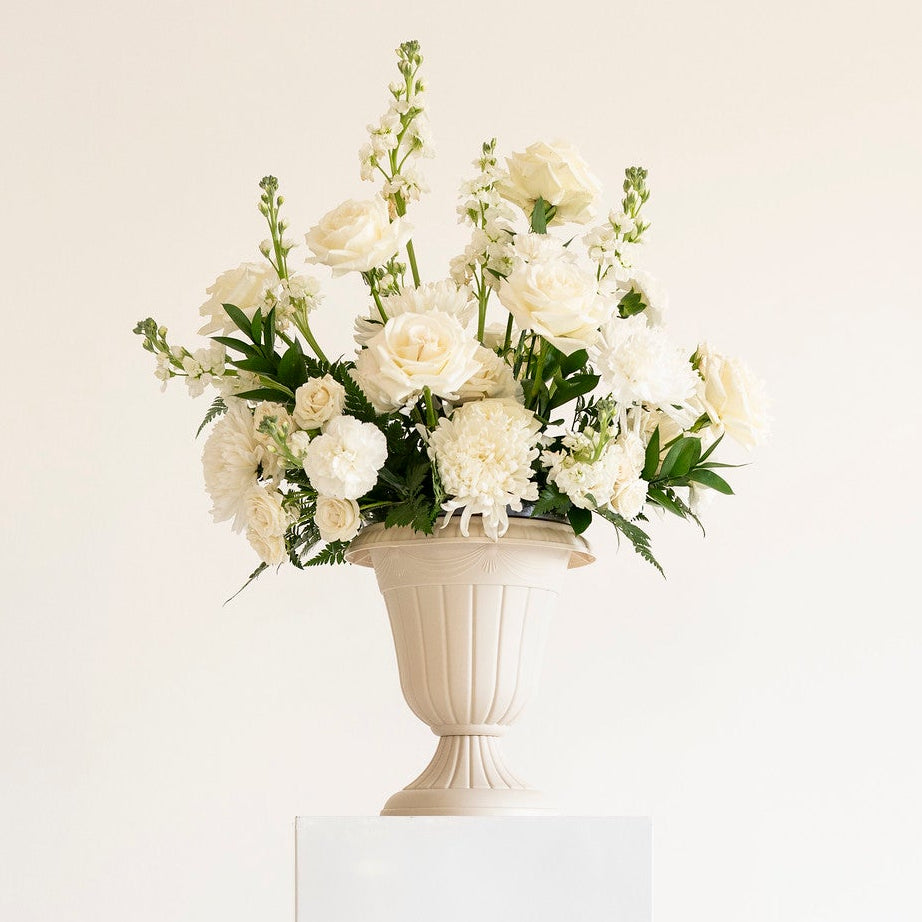 A white ceramic urn-shaped vase filled with white roses and green foliage, displayed on a white rectangular pedestal against a plain off-white background.
