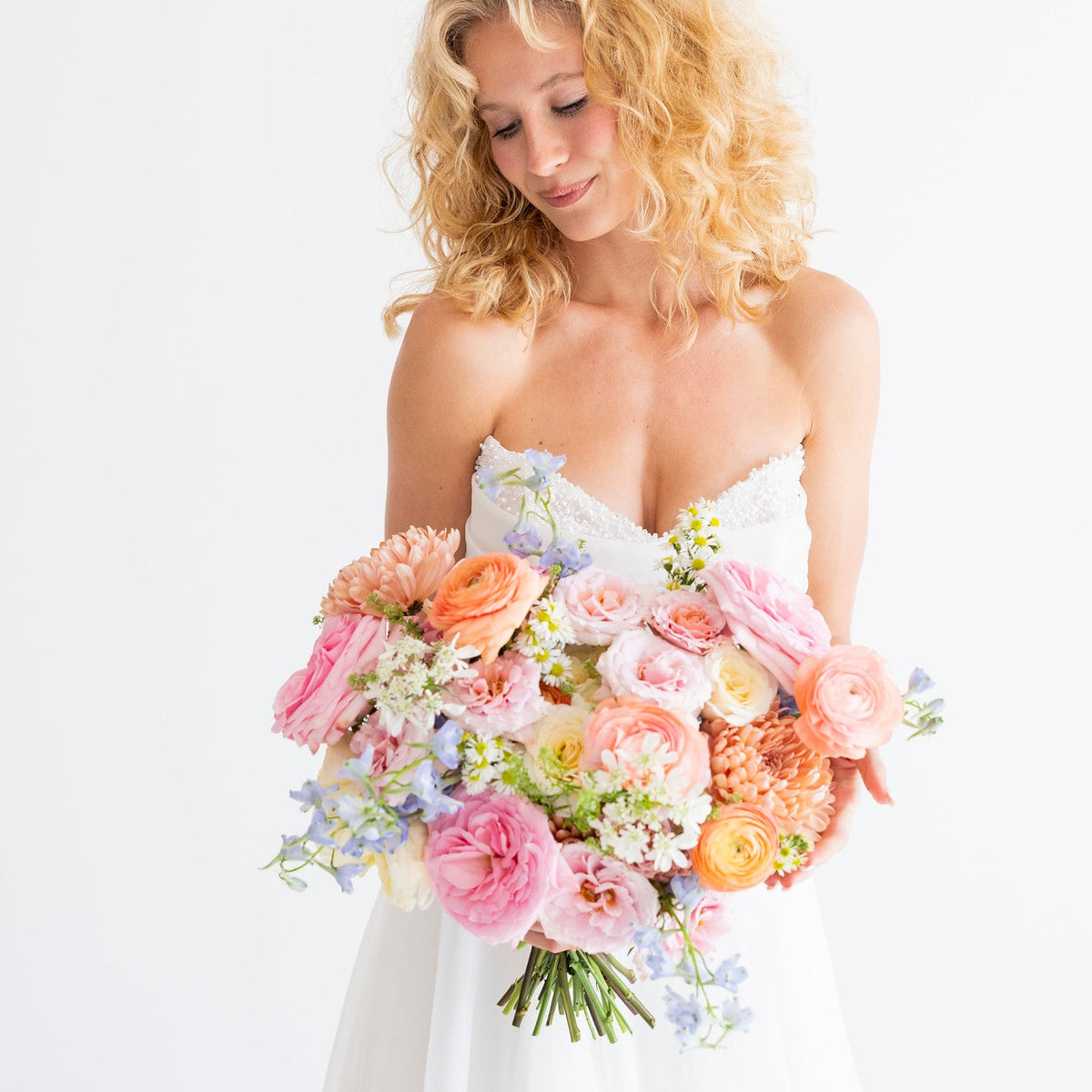 A blonde bride in a strapless white wedding gown holds a large pastel bouquet of pink, peach, orange, and light blue flowers against a clean white background.
