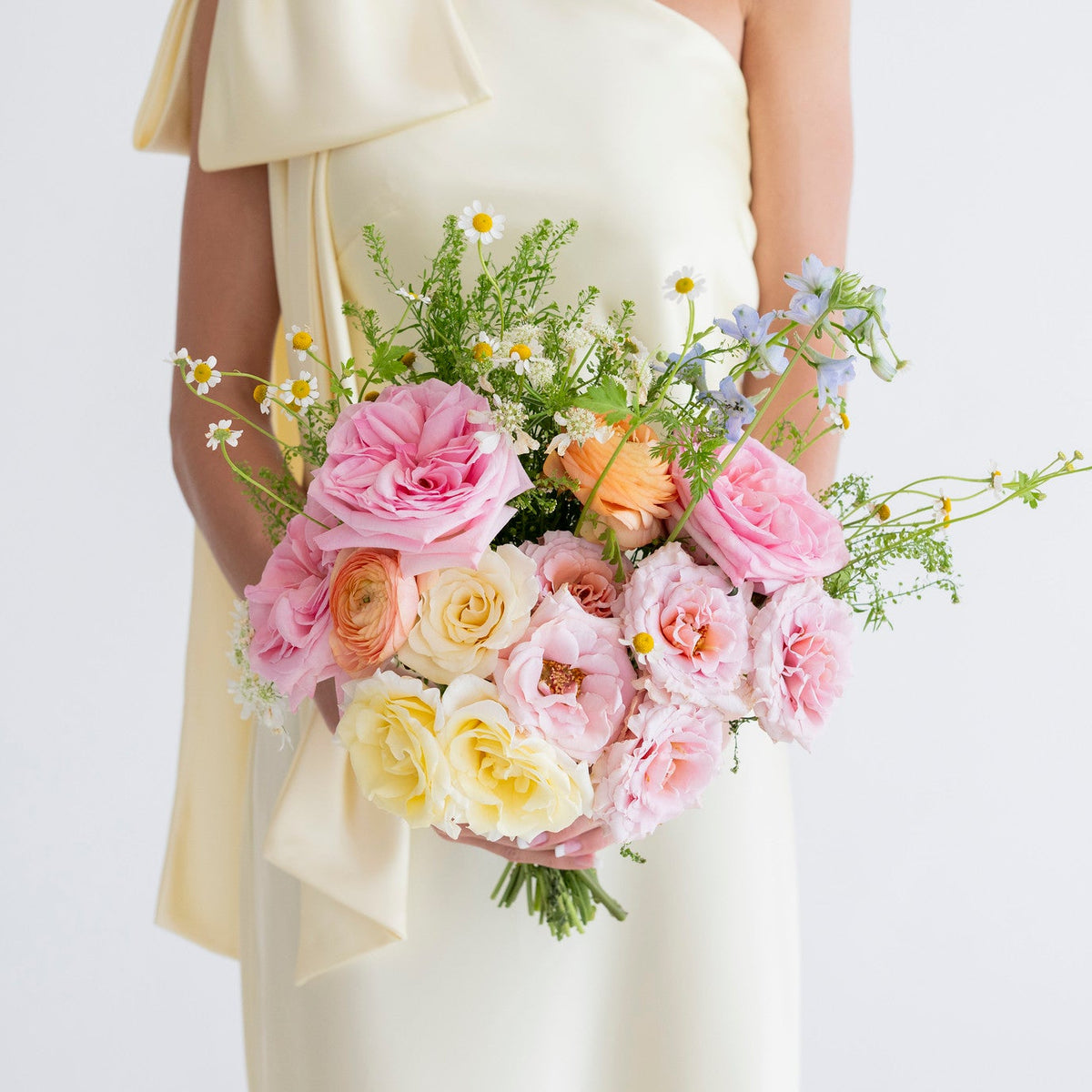 A person in a cream one-shoulder dress holding a pastel flower bouquet with pink, peach, and yellow roses, green foliage, and small white and blue wildflowers against a plain light background.
