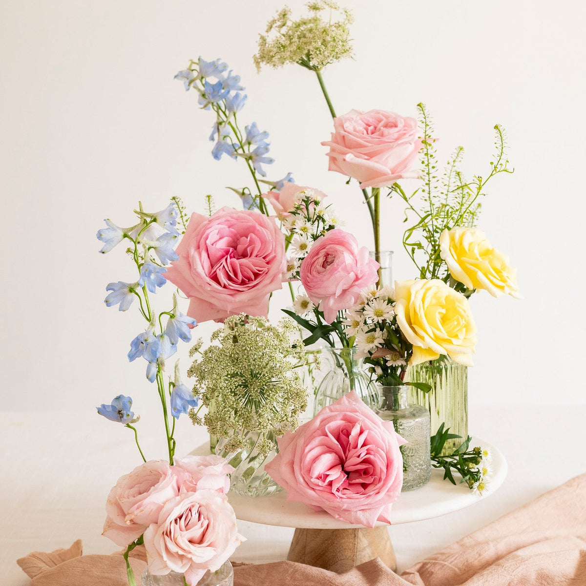 A floral arrangement of pink and yellow roses with blue delphiniums and green foliage in clear glass vases, styled on a draped blush linen cloth against a soft cream background.