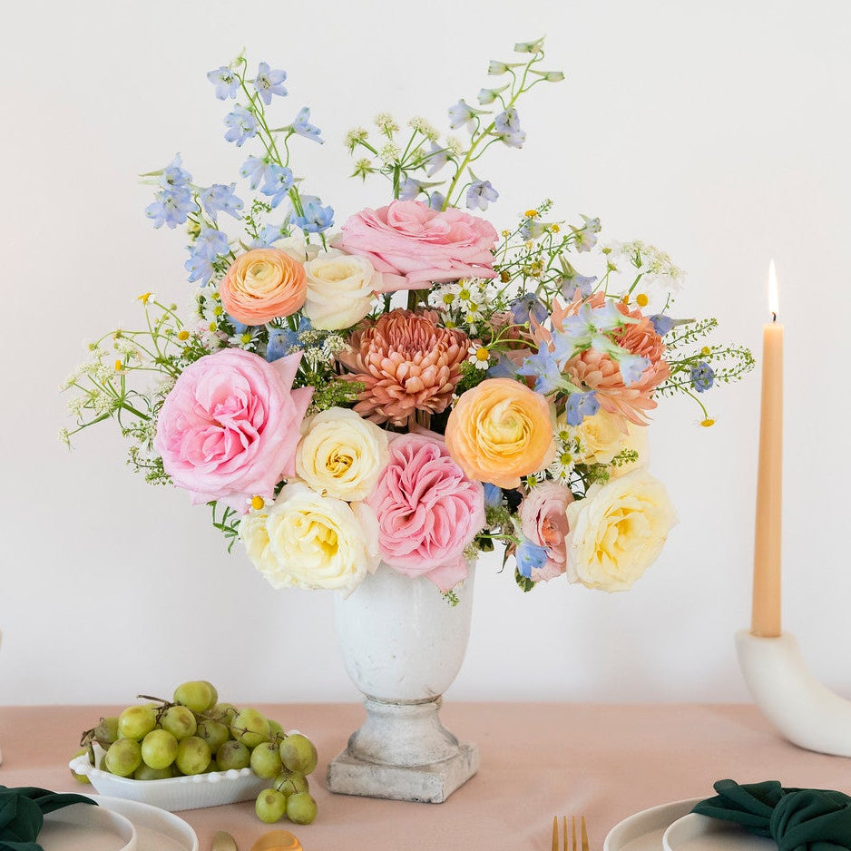 A styled table setting with a large floral centerpiece in a white urn, two place settings with white plates, gold cutlery, dark green napkins, a bowl of green grapes, and two tall lit taper candles on a blush pink tablecloth against a white wall.